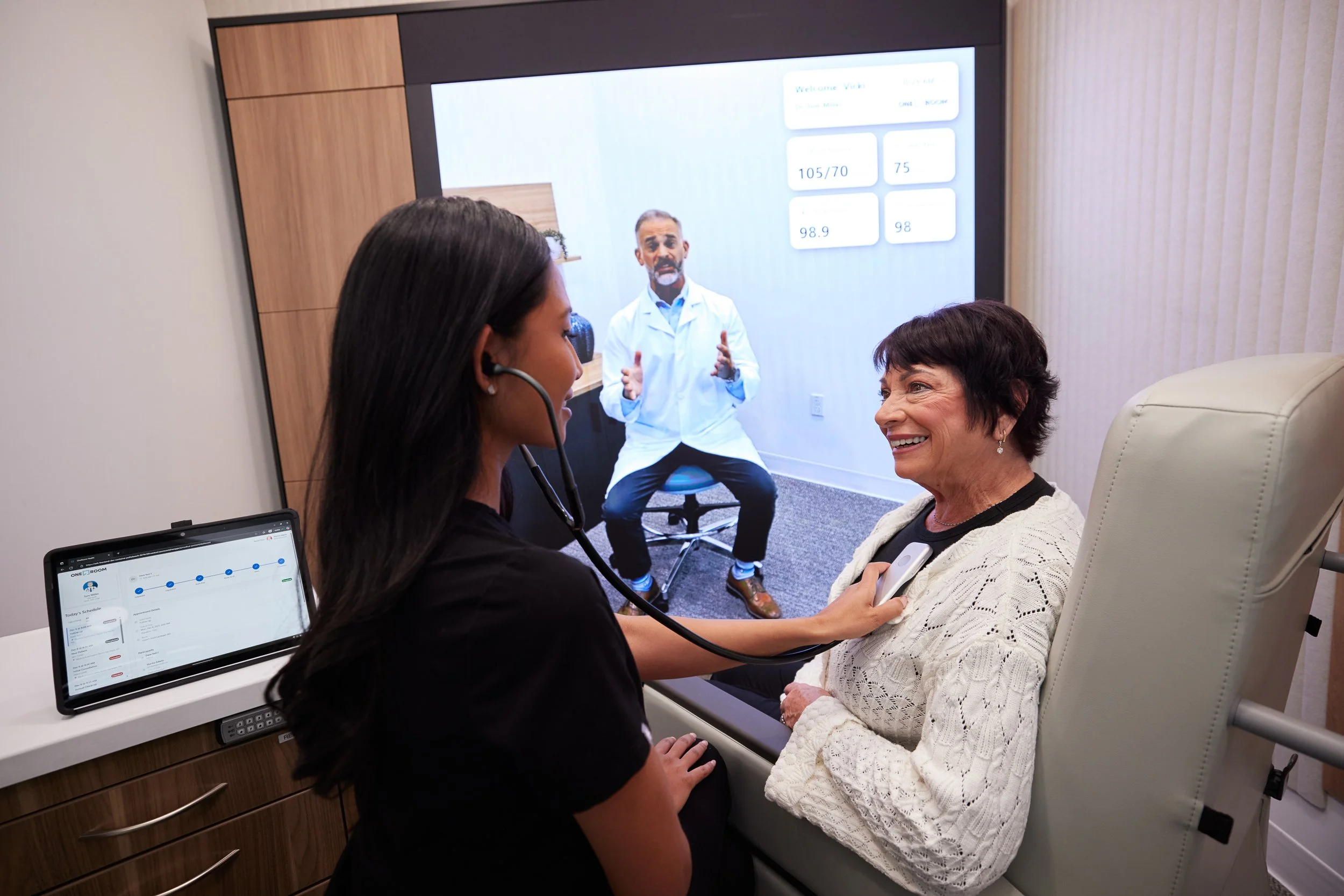 A healthcare professional using a stethoscope on an elderly woman in a medical examination room, with a large screen in the background showing a doctor on a video call.