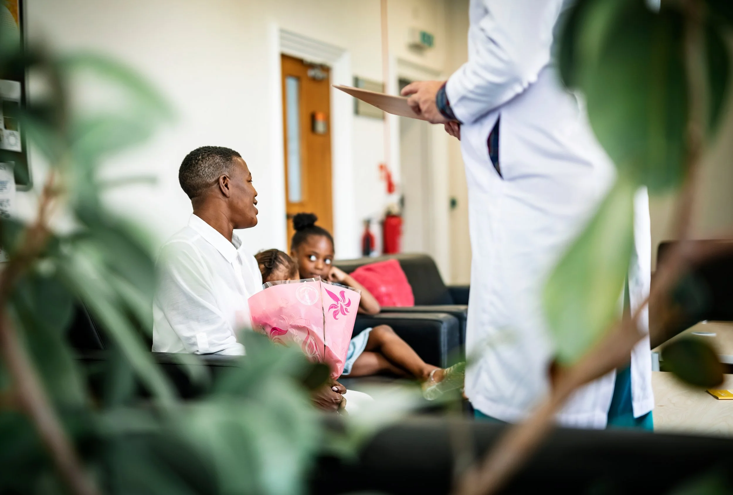 A man holding a bouquet of pink flowers sits on a bench and talks to a healthcare worker in a white coat. Two young girls are sitting nearby, one with her head resting on the other's shoulder, in a waiting room.