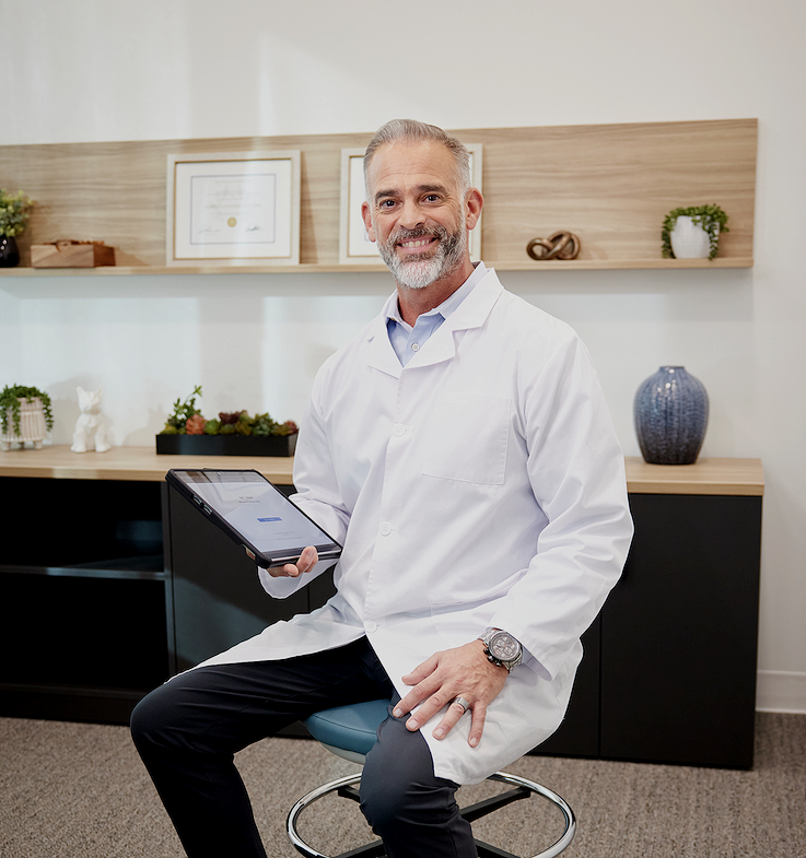 A cheerful middle-aged male doctor with gray hair and a beard, wearing a white coat, sitting on a stool in an office, holding a tablet.