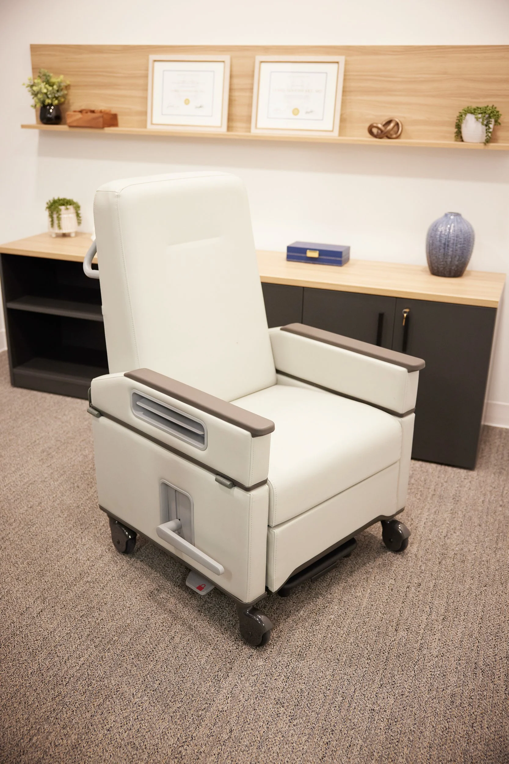 A white medical recliner chair with armrests and wheels in an office or clinic room, with a wooden shelf, framed certificates, decorative items, and plants on the wall behind.
