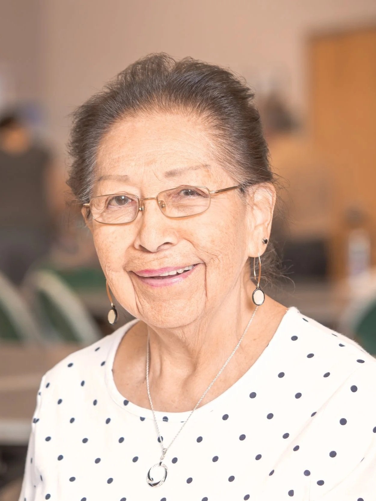 Smiling elderly woman with glasses, earrings, and necklace, wearing a white shirt with black polka dots, indoors.
