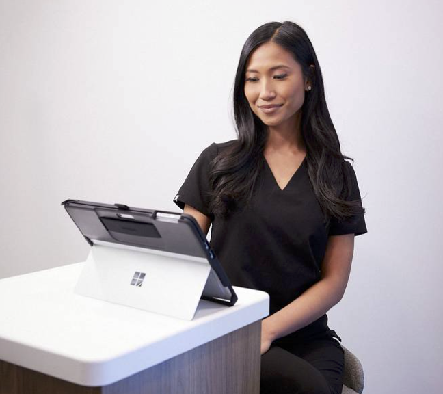 A woman in black scrubs sitting at a white table, looking at a Microsoft Surface device.