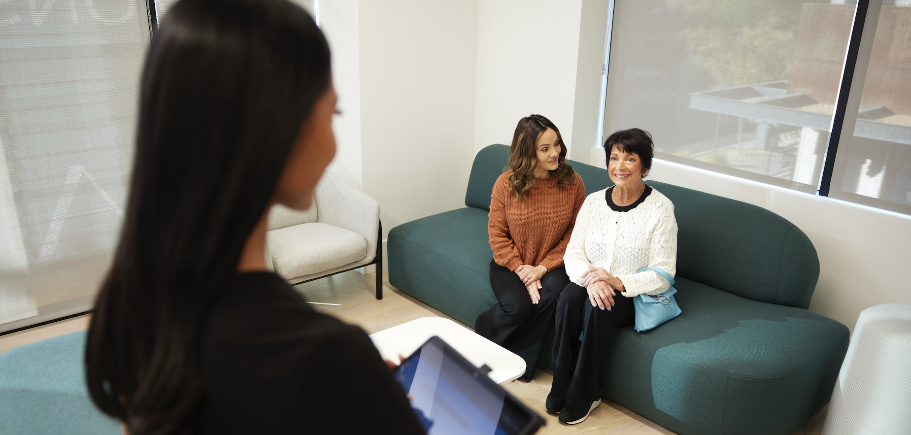 Two women sitting on a green sofa having a conversation, with a woman blurred in the foreground holding a tablet, in a bright room with large windows.