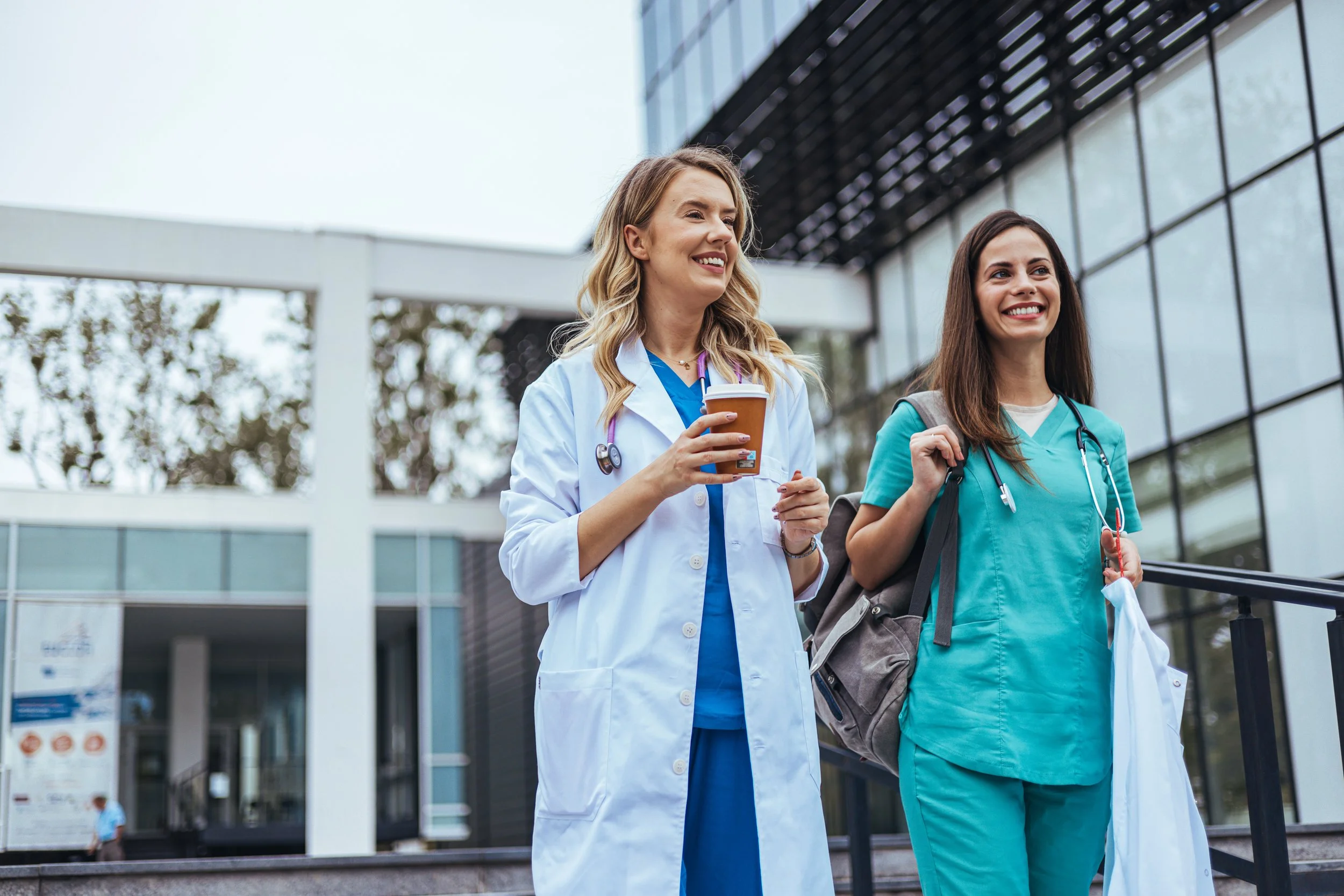 Two female healthcare professionals walking outside a modern medical facility; one in a white lab coat holding a coffee cup, the other in teal scrubs with a backpack and face mask.