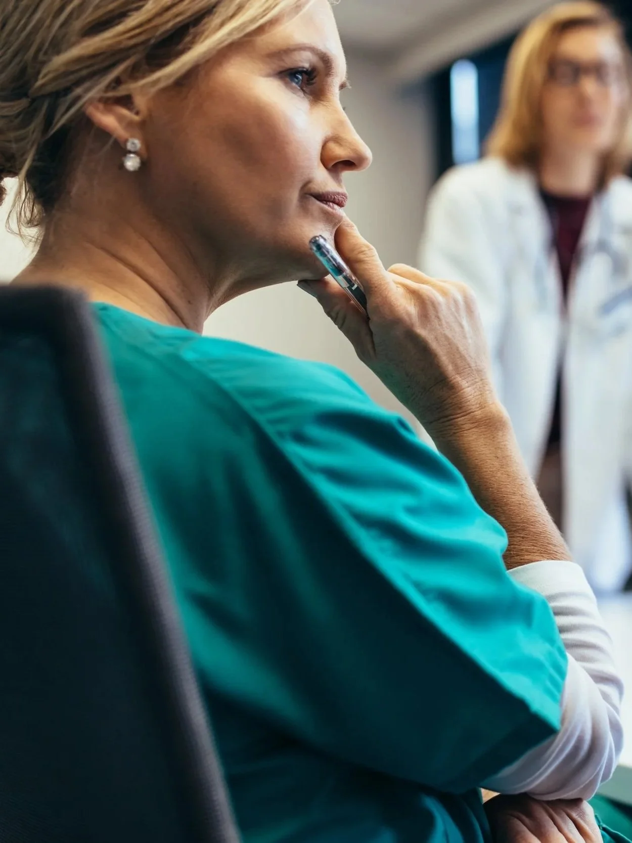 A woman in scrubs sitting with her hand on her chin, focused and thoughtful, with a healthcare worker in a white coat in the background.