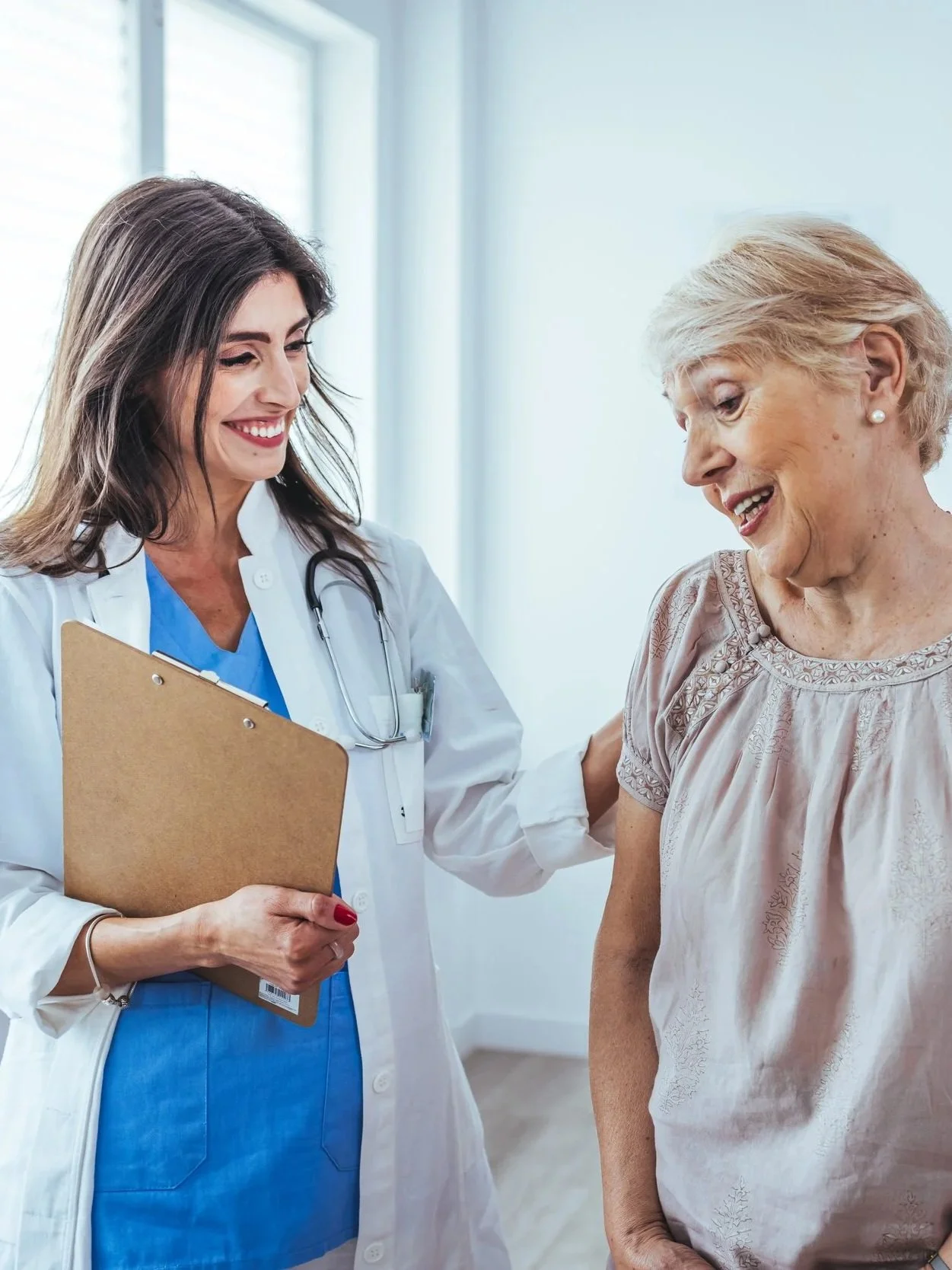 A healthcare professional, smiling and holding a clipboard, interacts with an elderly woman in a clinical setting.