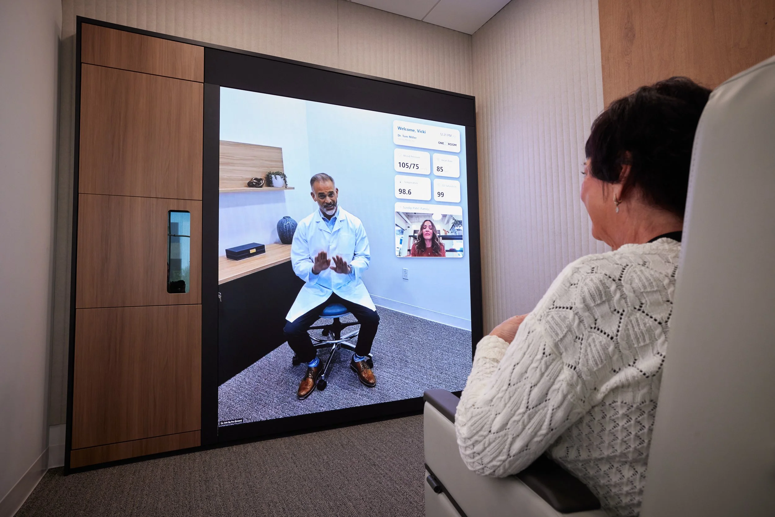 A woman seated in a medical or consultation room looking at a large screen showing a virtual doctor meeting with a male doctor in a white coat, with digital health data displayed on the screen.