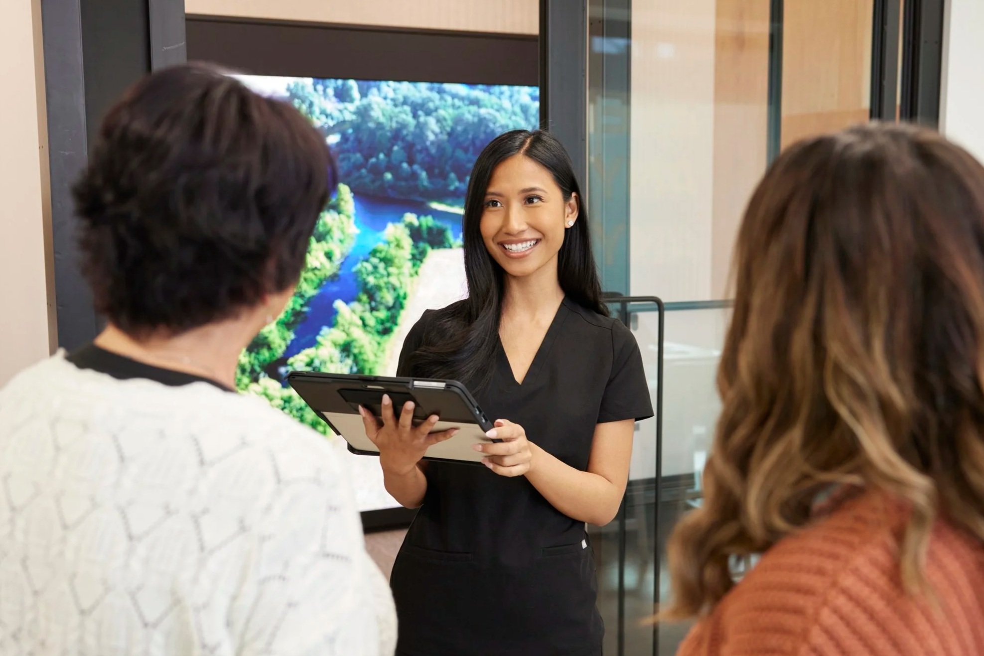 A smiling woman in black scrubs holding a tablet, talking to two women with wavy hair in a professional setting, with a large screen displaying a nature scene of a river and trees in the background.