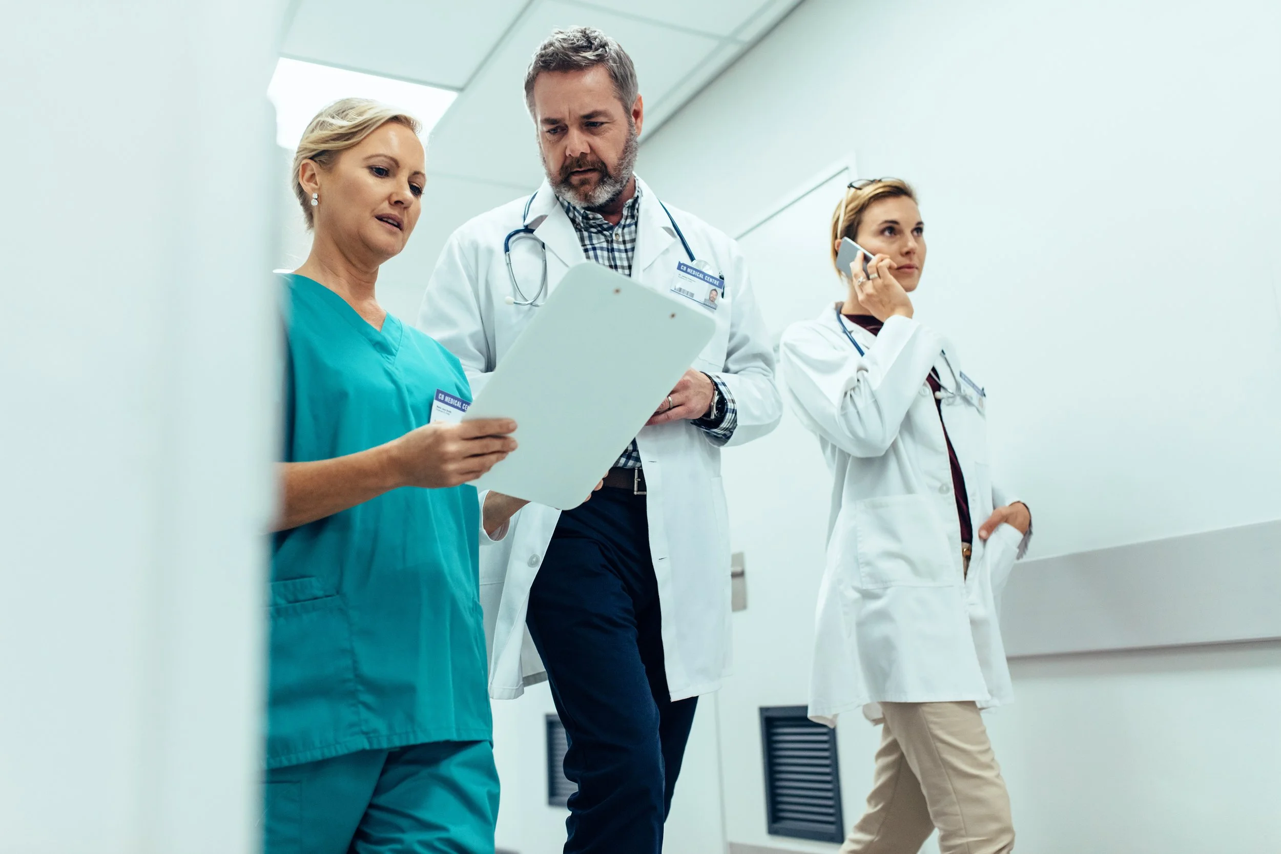 Three medical professionals, including two women and one man, review documents and talk on a cell phone in a hospital corridor.