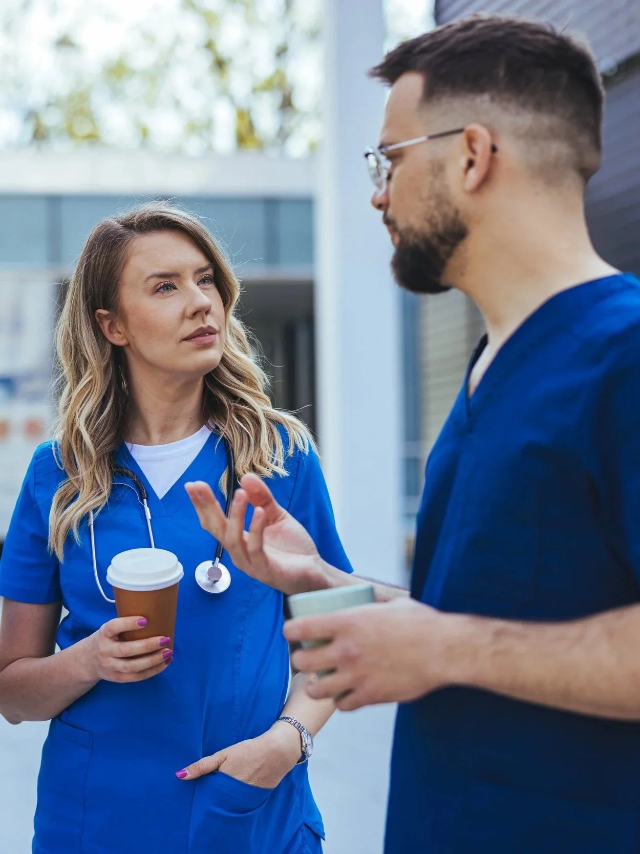 Two healthcare professionals, wearing blue scrubs, are having a conversation outdoors. One woman is holding a coffee cup, and a man is explaining something using hand gestures. The woman has a stethoscope draped around her neck.