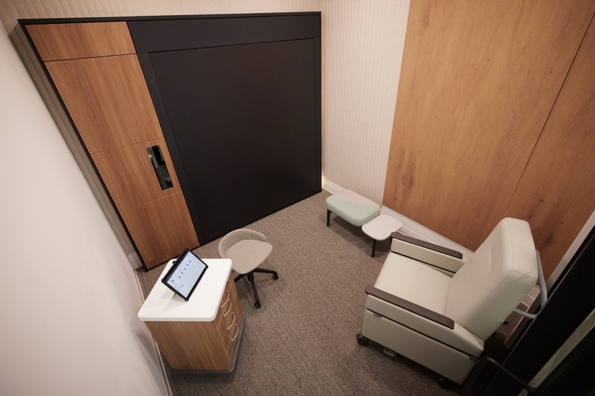 An office or therapy room with a beige reclining chair, small tables, a small gray upholstered stool, a chair, a white dresser with a tablet on top, a large black and brown wardrobe, and beige walls and carpet.