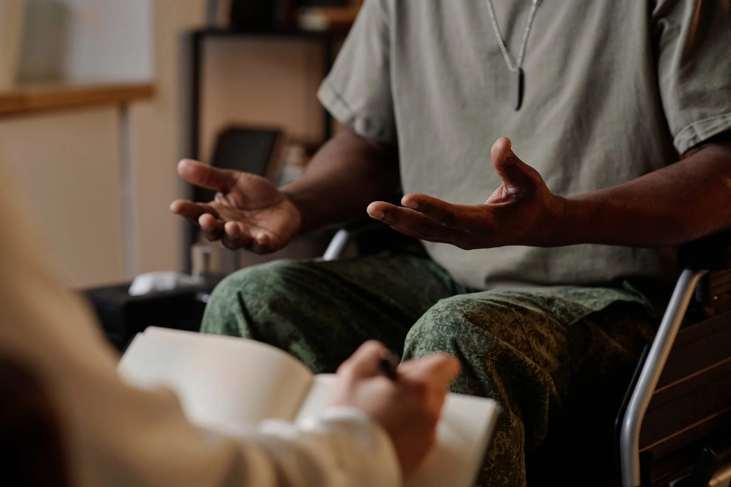 A person in military camouflage pants and a gray t-shirt gesturing with their hands while speaking to a person holding a notebook and pen.