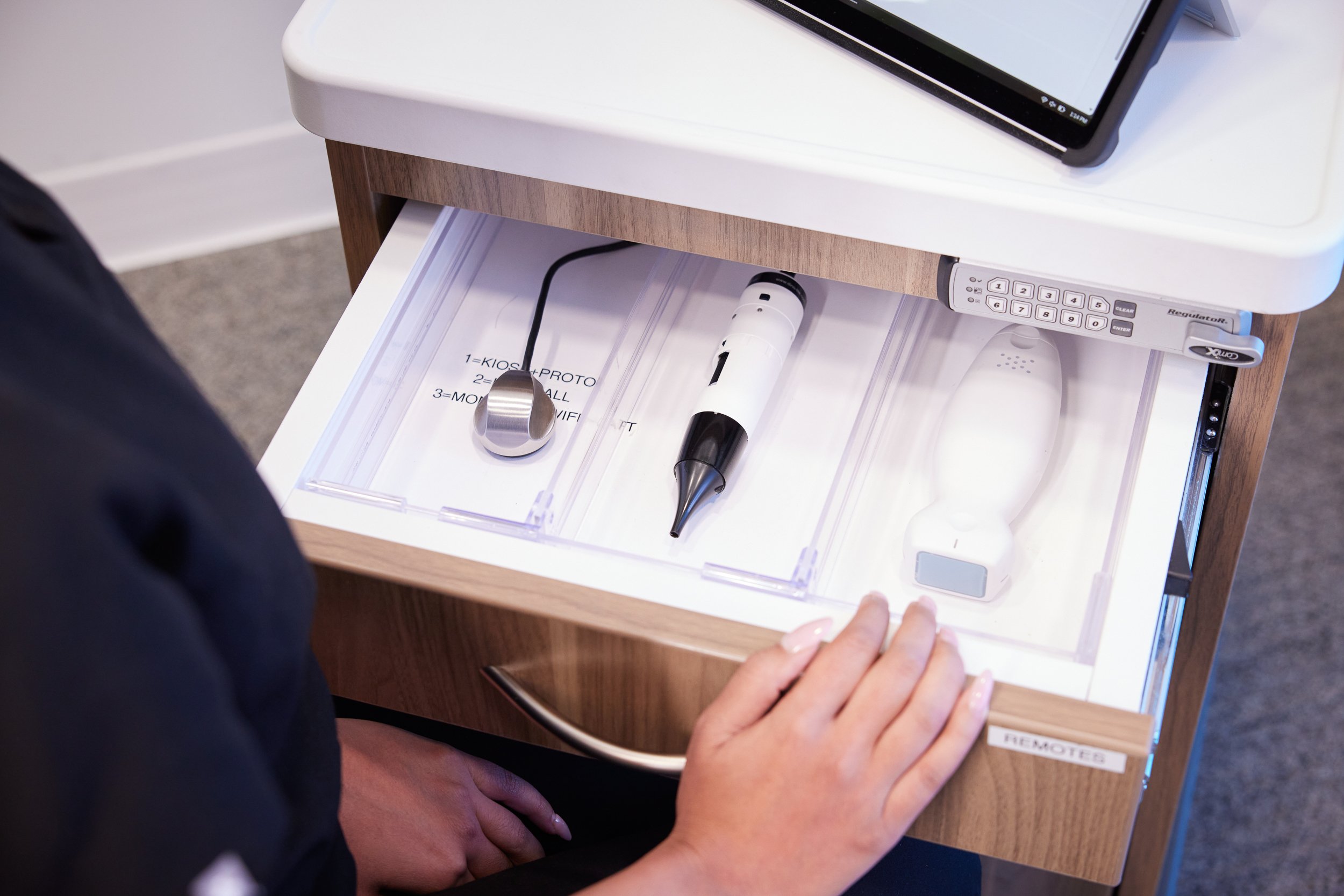 Open drawer with medical devices including a handheld otoscope, a reflex hammer, and a tuning fork, with a tablet on top of the drawer.
