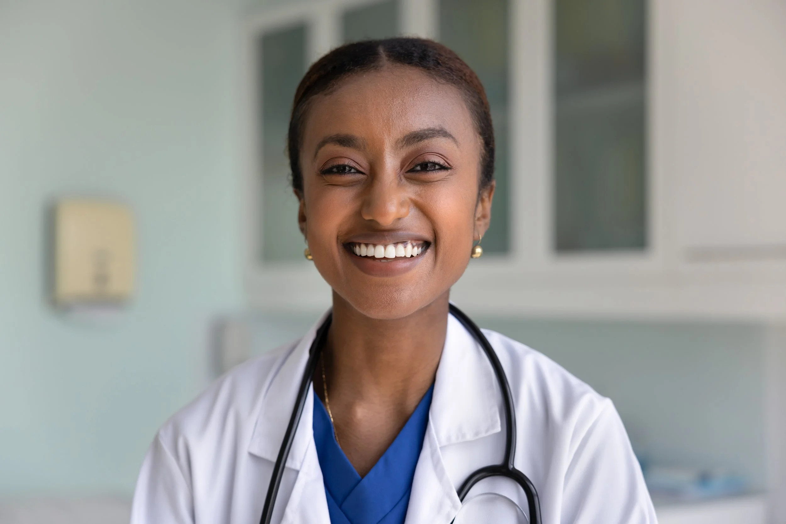 Smiling female doctor in white coat with stethoscope, indoors, in front of a window.