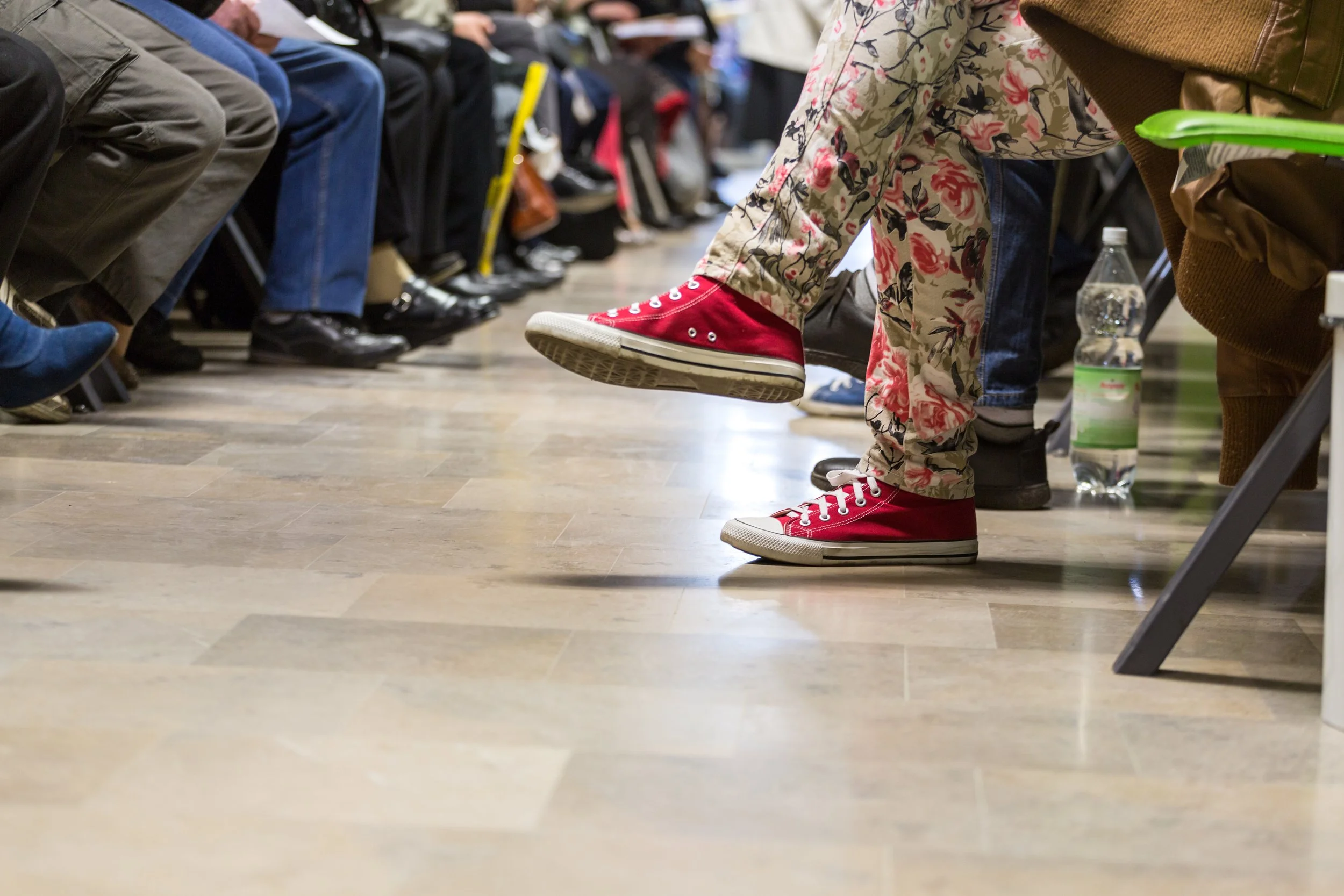 People sitting in rows of chairs, with one person in the foreground wearing bright red sneakers and floral-patterned pants, and a water bottle on the floor nearby.