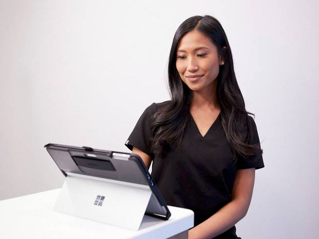 A woman with long black hair wearing a black shirt looking at a tablet on a white table against a plain white wall.