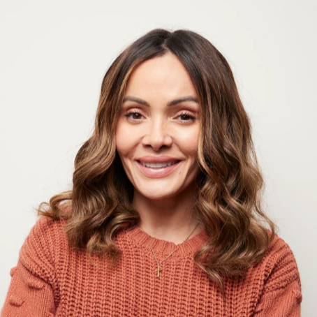 A woman with medium-length, wavy brown hair, smiling, wearing a rust-colored knit sweater, standing against a plain white background.