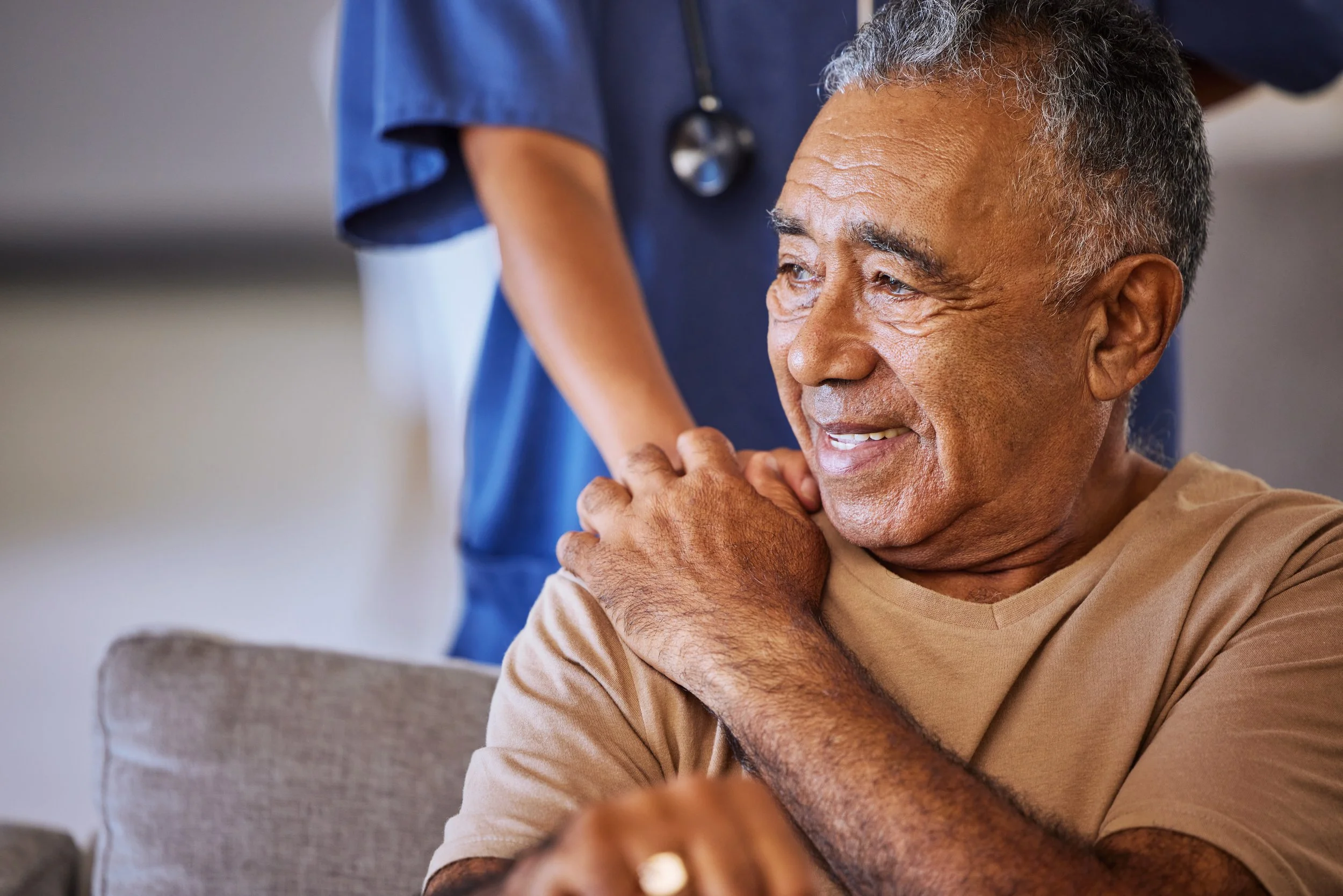 An elderly man smiling while touching his shoulder, with a healthcare professional in blue scrubs in the background.