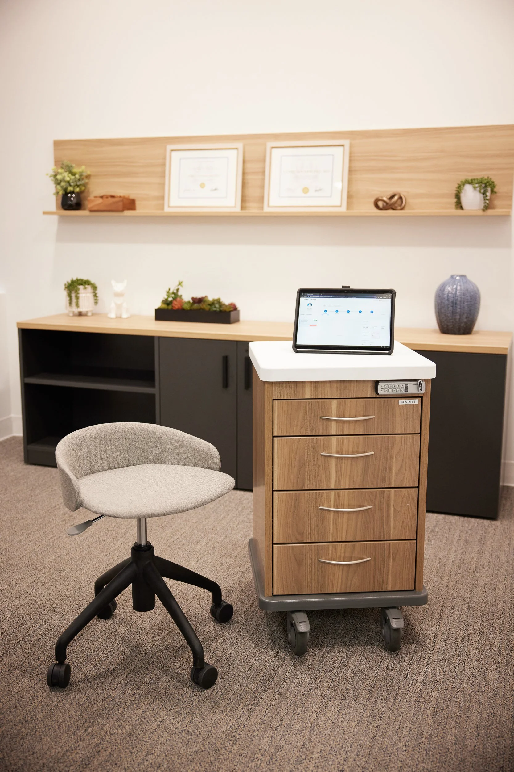 Office space featuring a beige rolling drawer cart with a tablet on top, a light gray office chair, and a dark cabinet with decorative plants, a cat figurine, and framed certificates on a shelf.