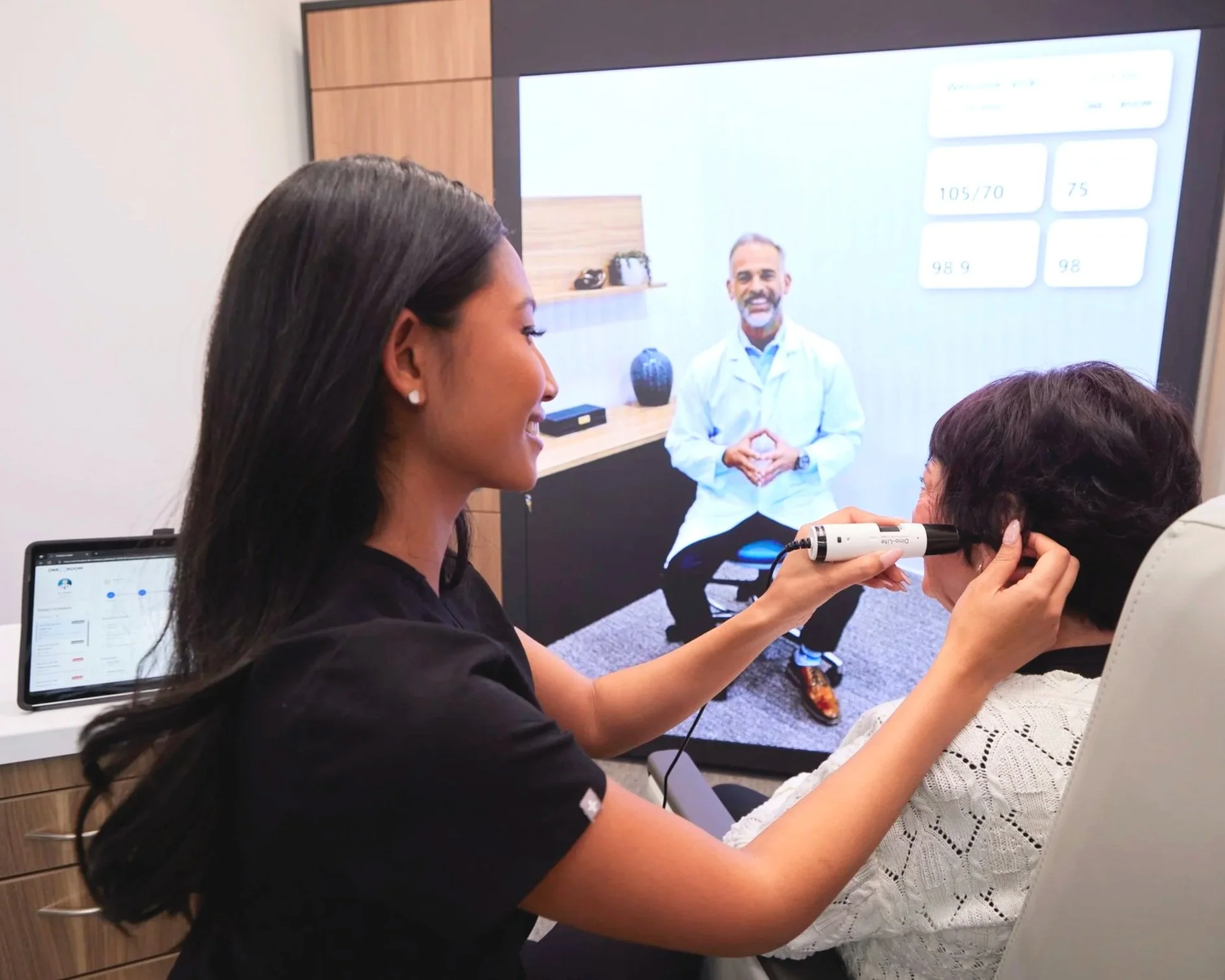 Medical professional using an otoscope to examine an elderly woman's ear during a telehealth consultation, with a large screen displaying a doctor in a white coat in the background.