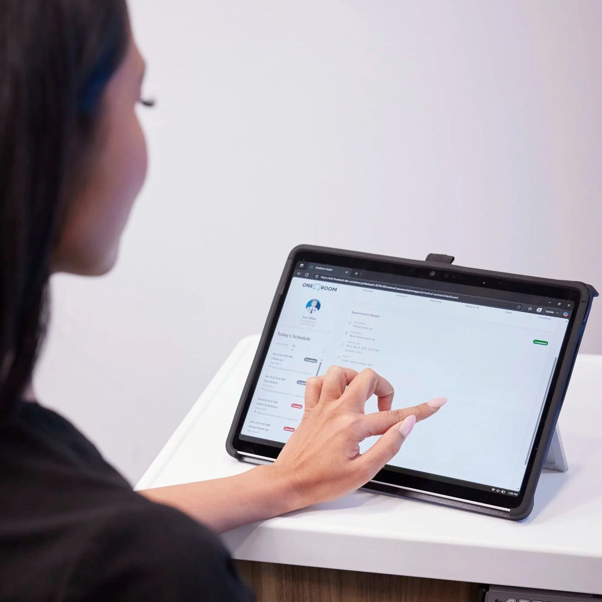 A woman using a tablet with a stylus, viewing a scheduling application on a white desk.