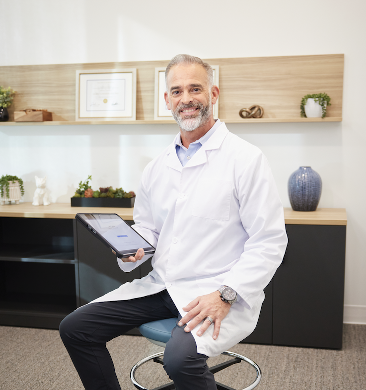 A doctor sitting on a stool in an office, holding a tablet, smiling at the camera, with framed certificates and decorative objects in the background.