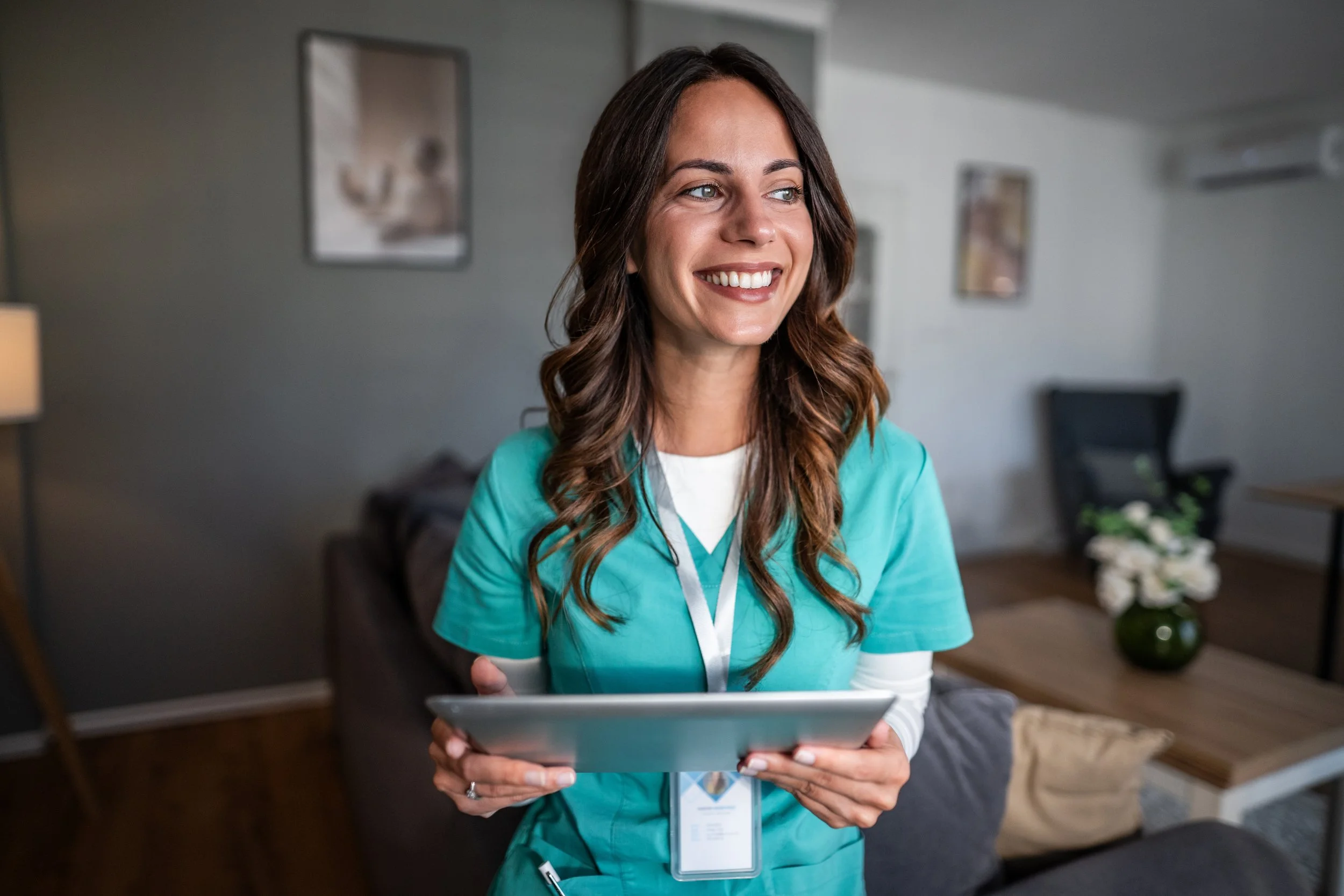 A smiling woman in medical scrubs holding a tablet, standing in a living room.