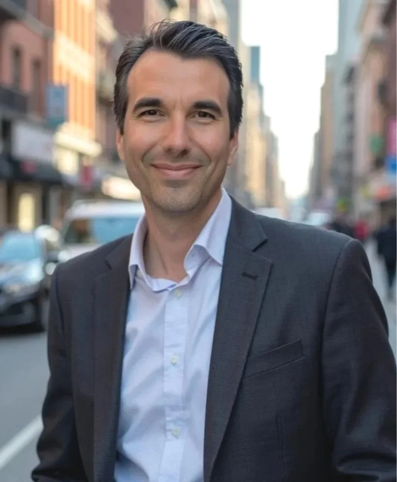 A smiling man in a dark suit and white shirt standing on a city street with buildings and cars in the background.
