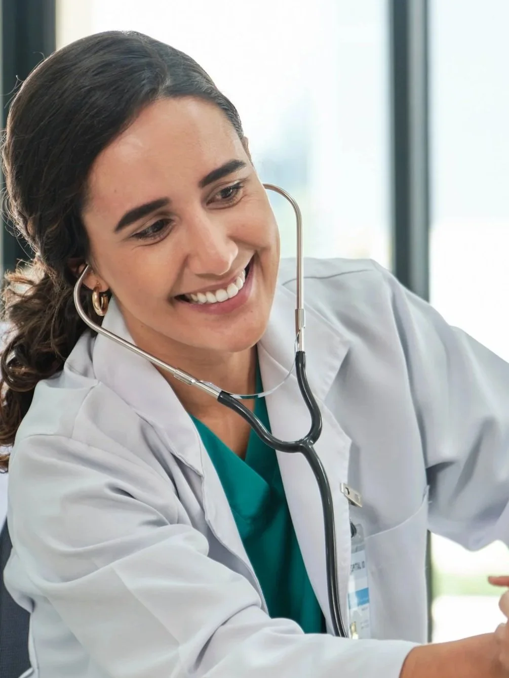 A smiling female doctor or nurse with a stethoscope around her neck, wearing a white coat and green scrubs, sitting at a desk in a bright medical office.