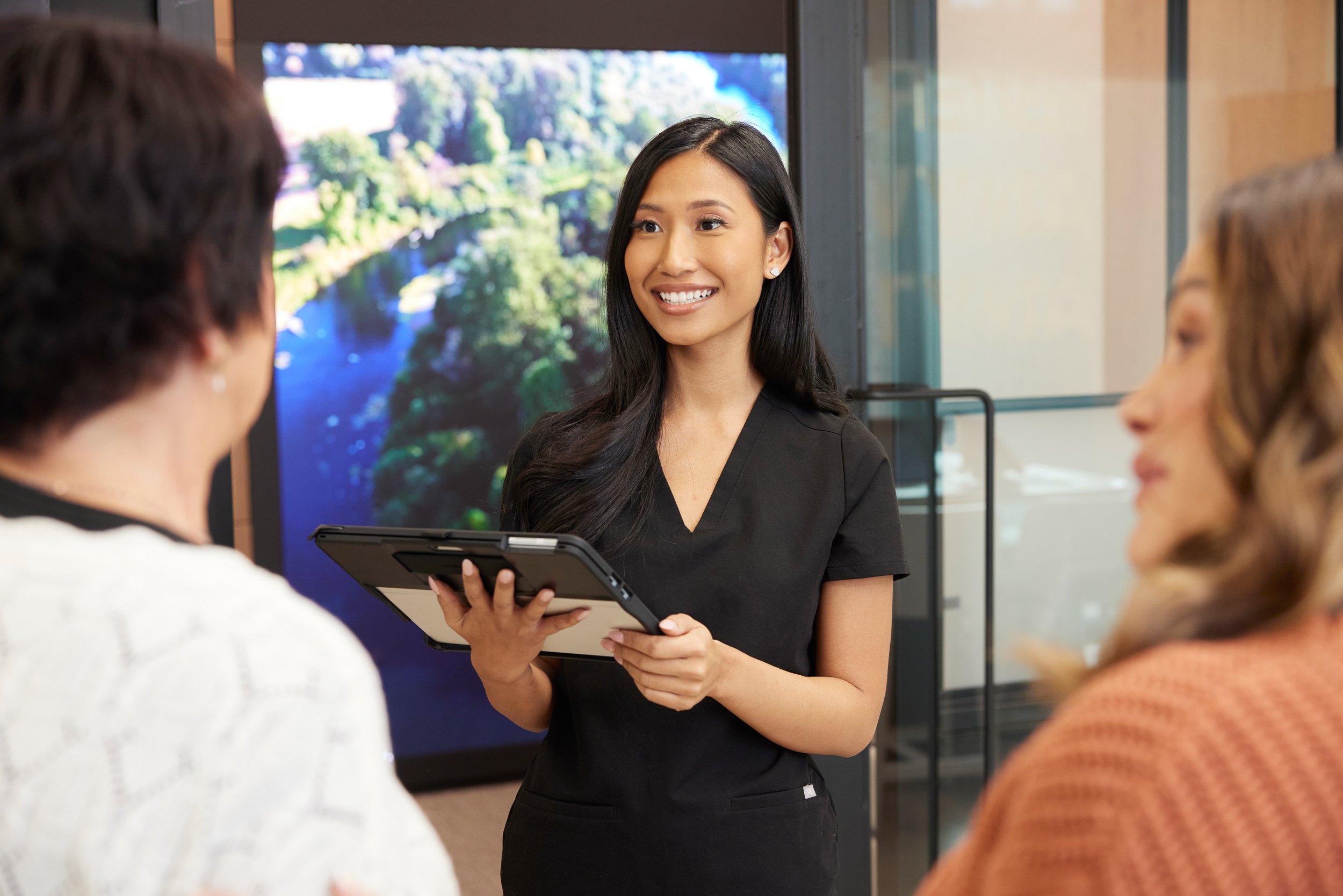 A woman with long dark hair, wearing a black uniform, smiling and holding a tablet, appears to be speaking to two women in a professional setting.