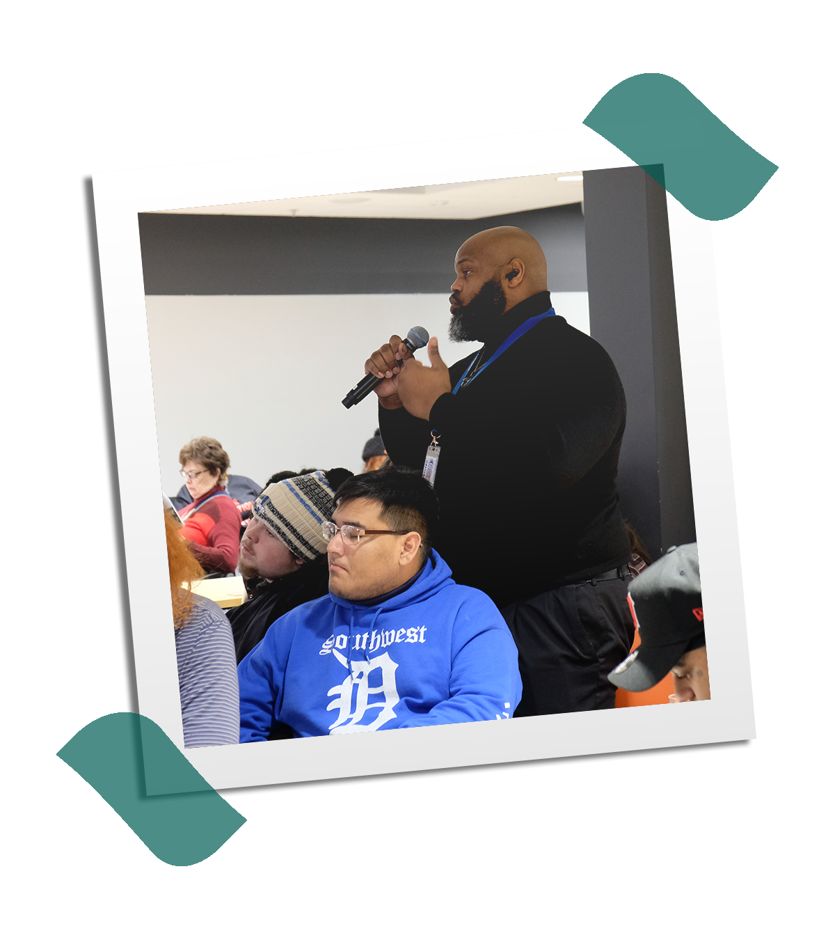 A man standing and speaking into a microphone during a conference or meeting, with several seated people listening, in an indoor setting.