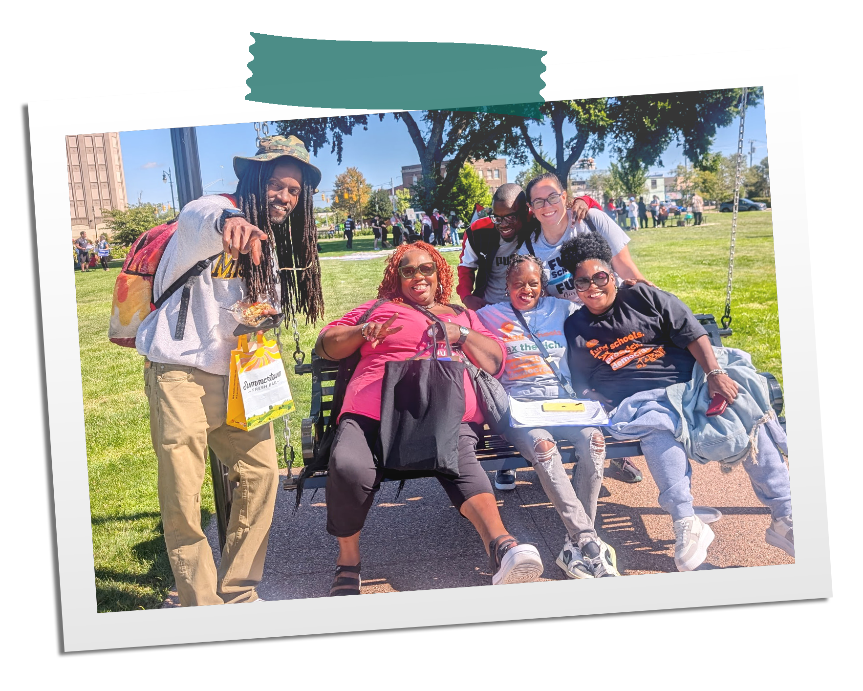 Group of people smiling and posing for a photo on a park bench on a sunny day with green grass and trees in the background.
