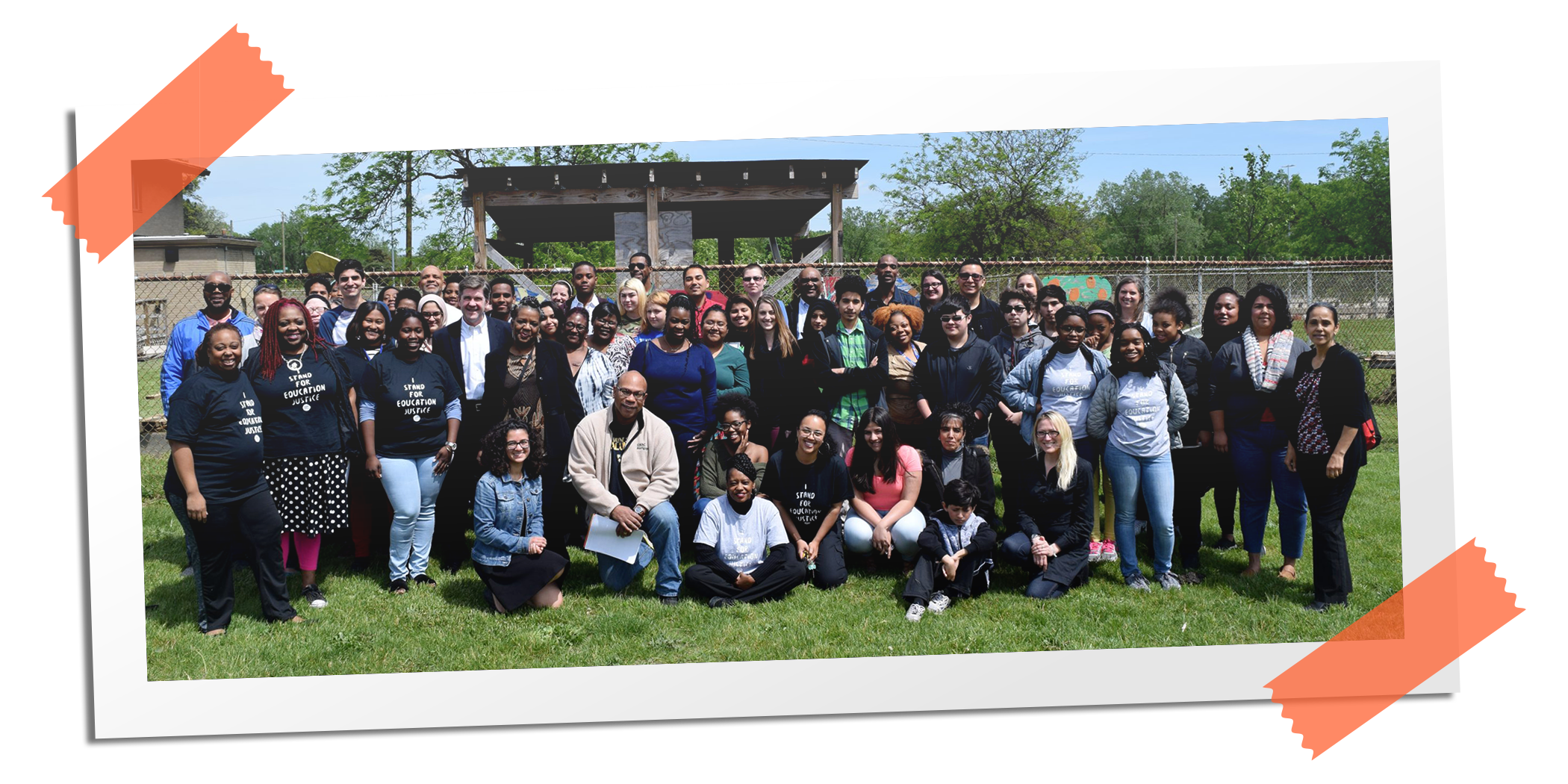 A large group of diverse people outdoors in a park, standing and sitting on the grass, with some wearing shirts that say 'Stand for Education Justice.' Background shows a chain-link fence and trees.