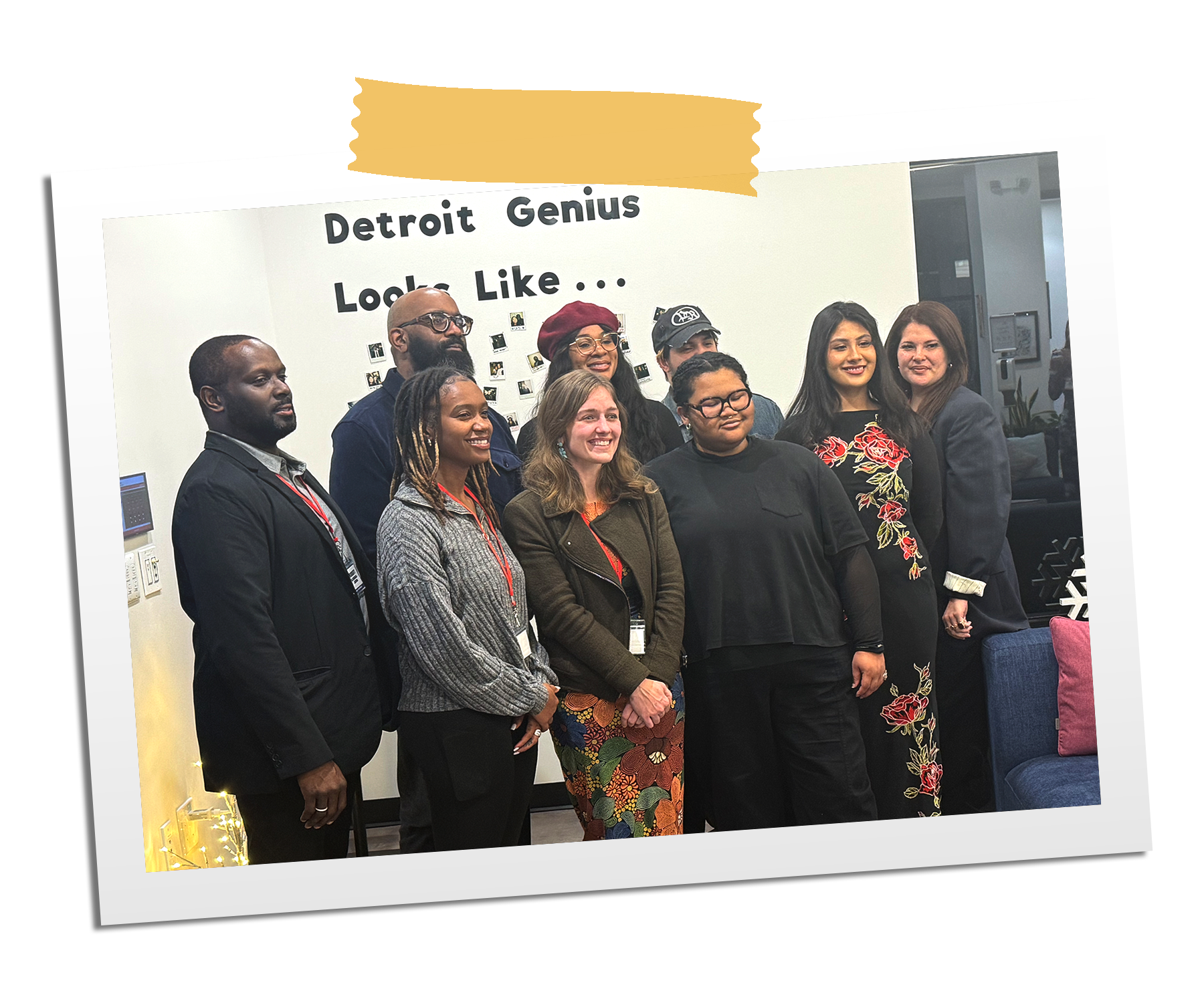 A group of nine diverse people standing indoors, smiling for a photo in front of a wall with the words "Detroit Genius Look Like..." on it.