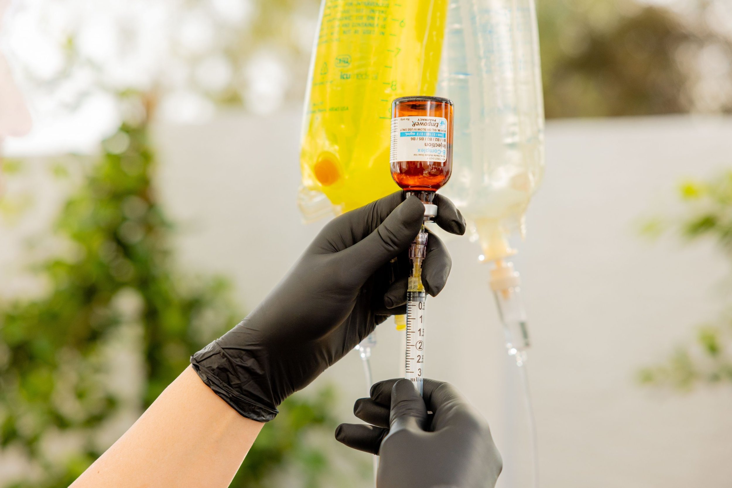 A person wearing black gloves fills a syringe with red medication from a small vial, with IV bags in the background.