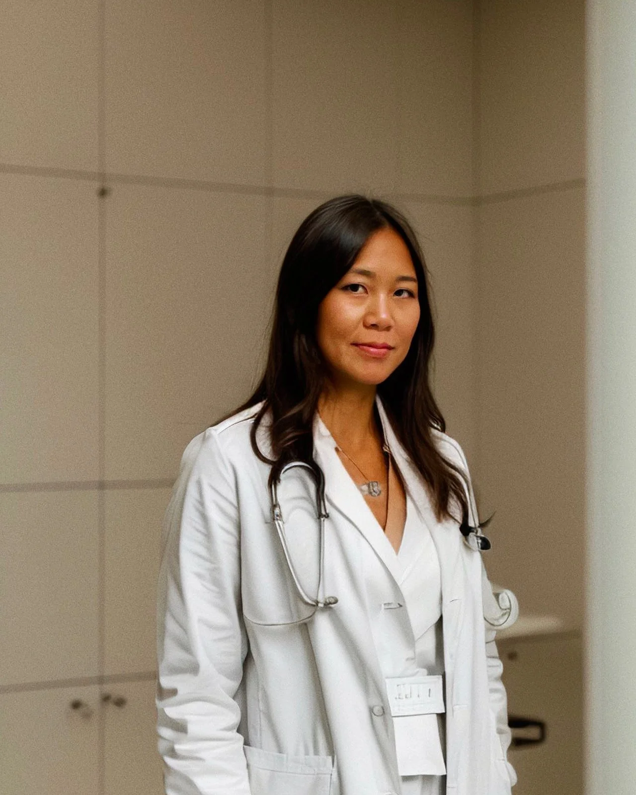 A female doctor in a white coat with a stethoscope around her neck standing in a hospital corridor.