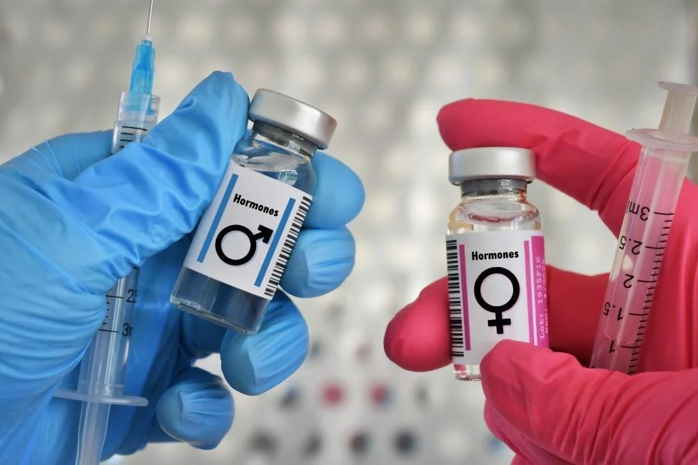 Two hands wearing blue and pink gloves hold small glass vials labeled with male and female symbols and the word 'Hormones'. One vial has a syringe nearby.