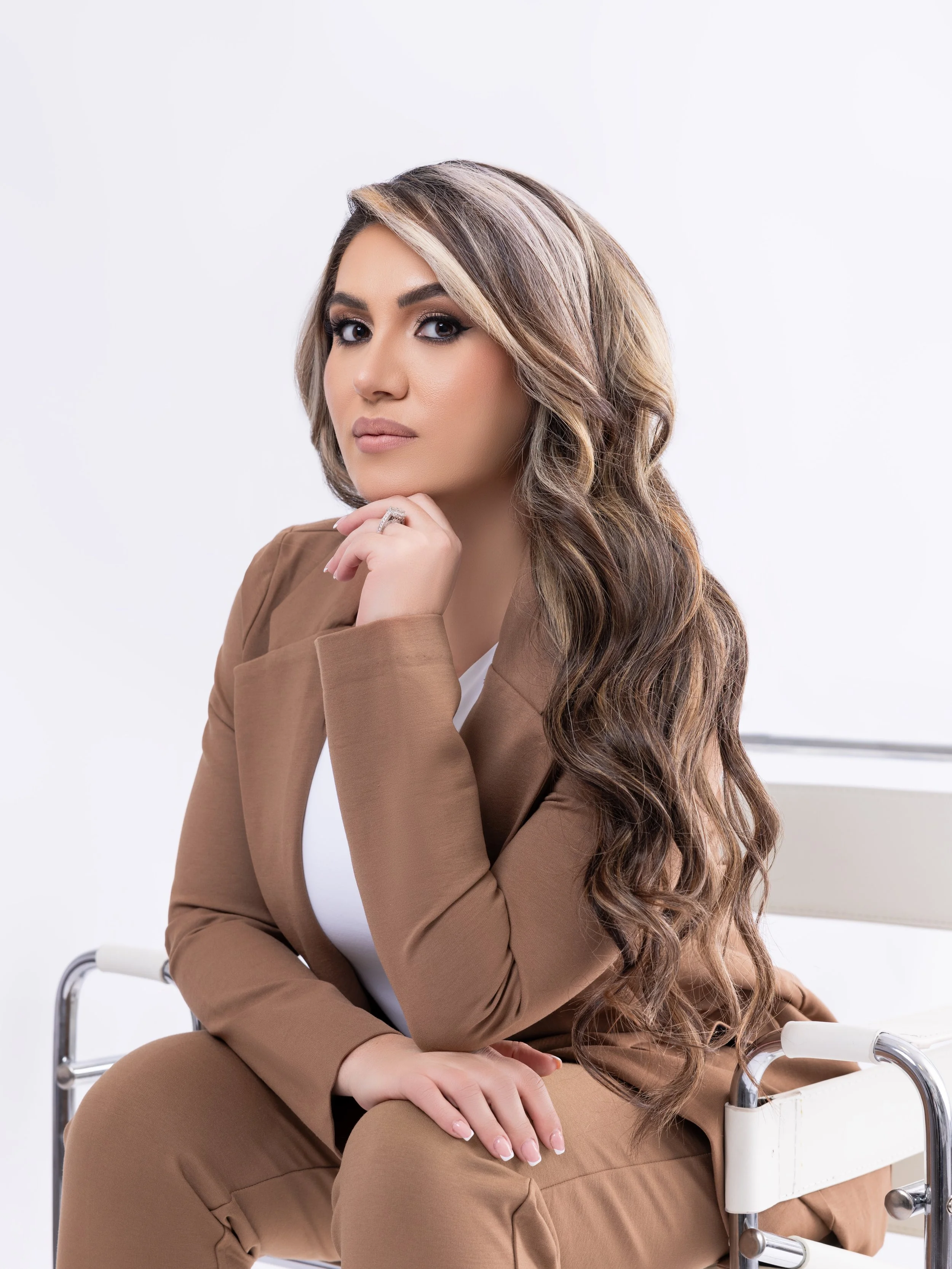 A woman with long, wavy brown hair and light makeup, wearing a brown blazer and pants, sitting on a white chair in a professional setting, with a neutral background.