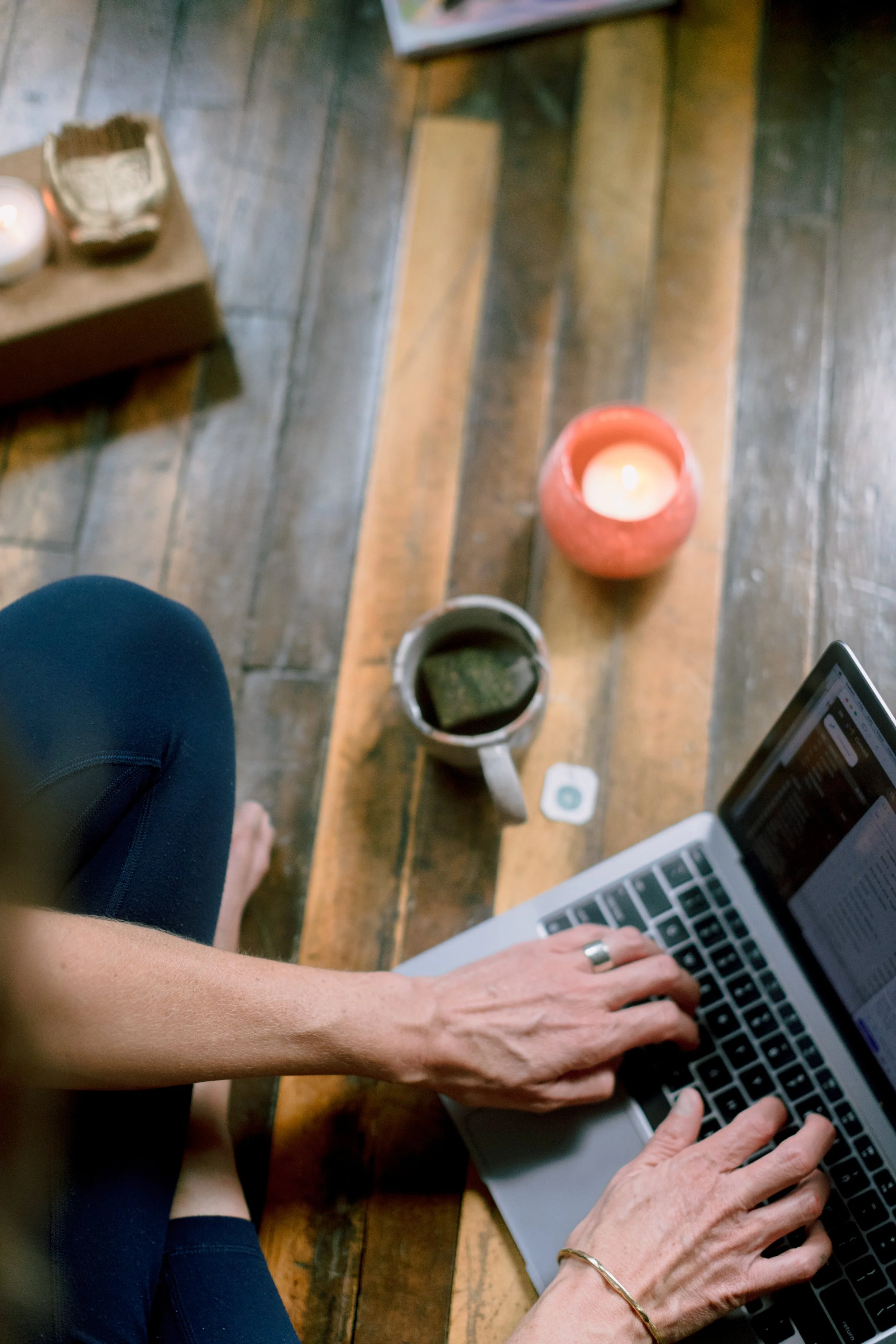 A person typing on a laptop keyboard, sitting at a wooden table with cups, a candle, and other objects.