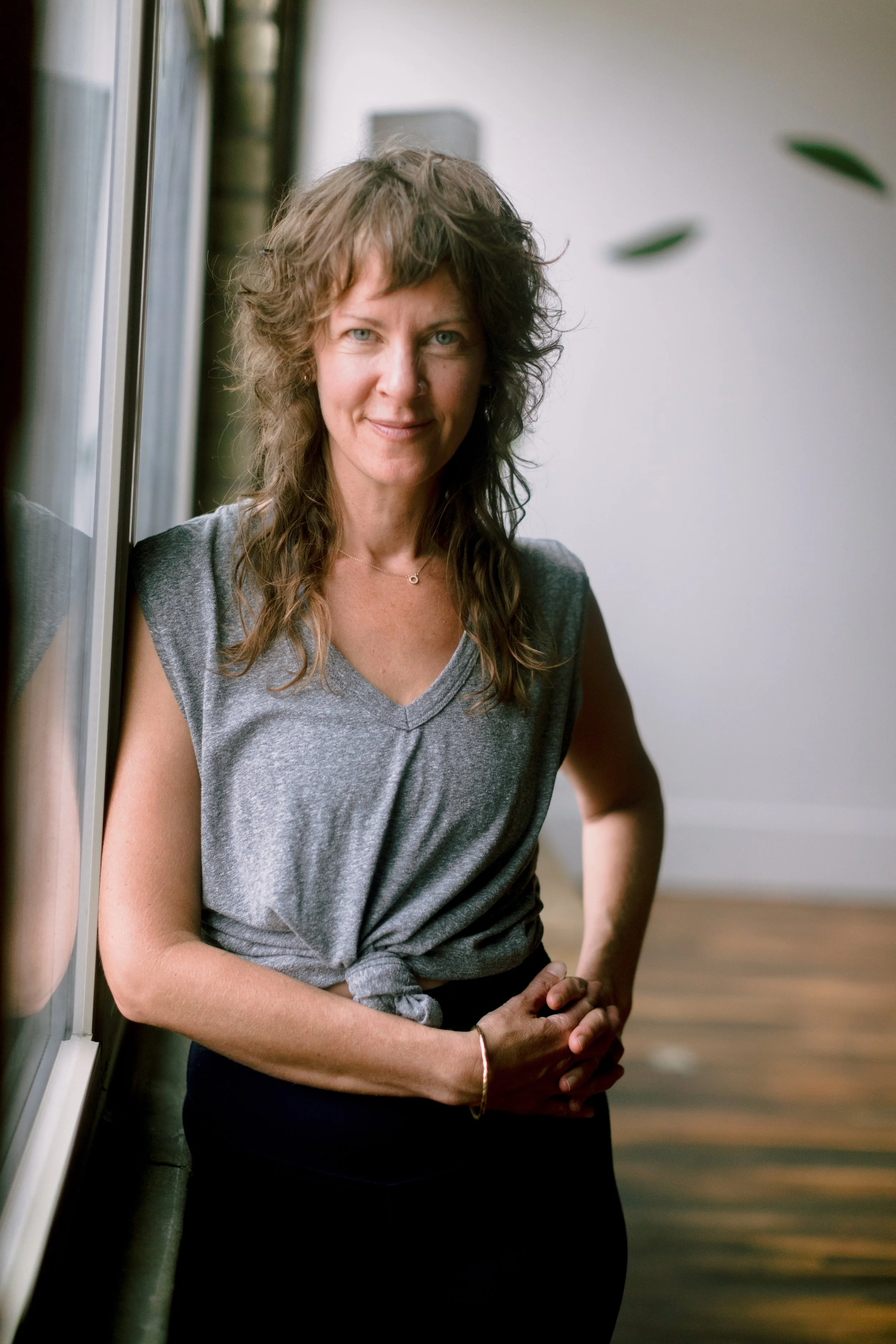 A woman with wavy, shoulder-length brown hair standing by a window, wearing a grey sleeveless top, with a gentle smile.