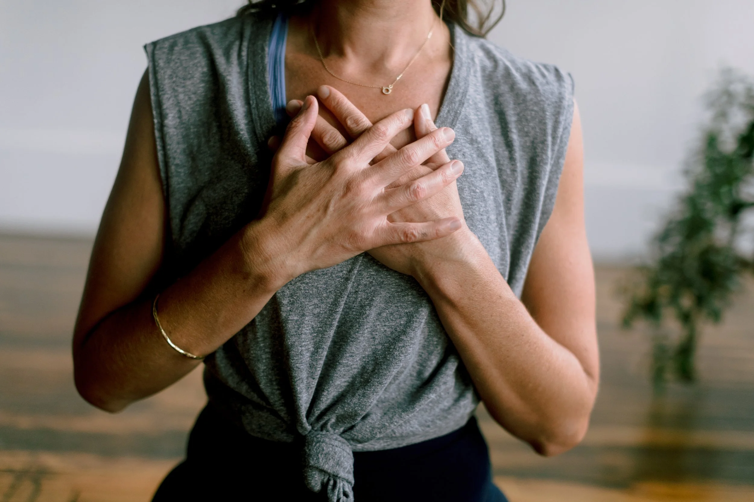 A woman in a gray sleeveless top holding her heart with both hands, possibly experiencing connection and ease.