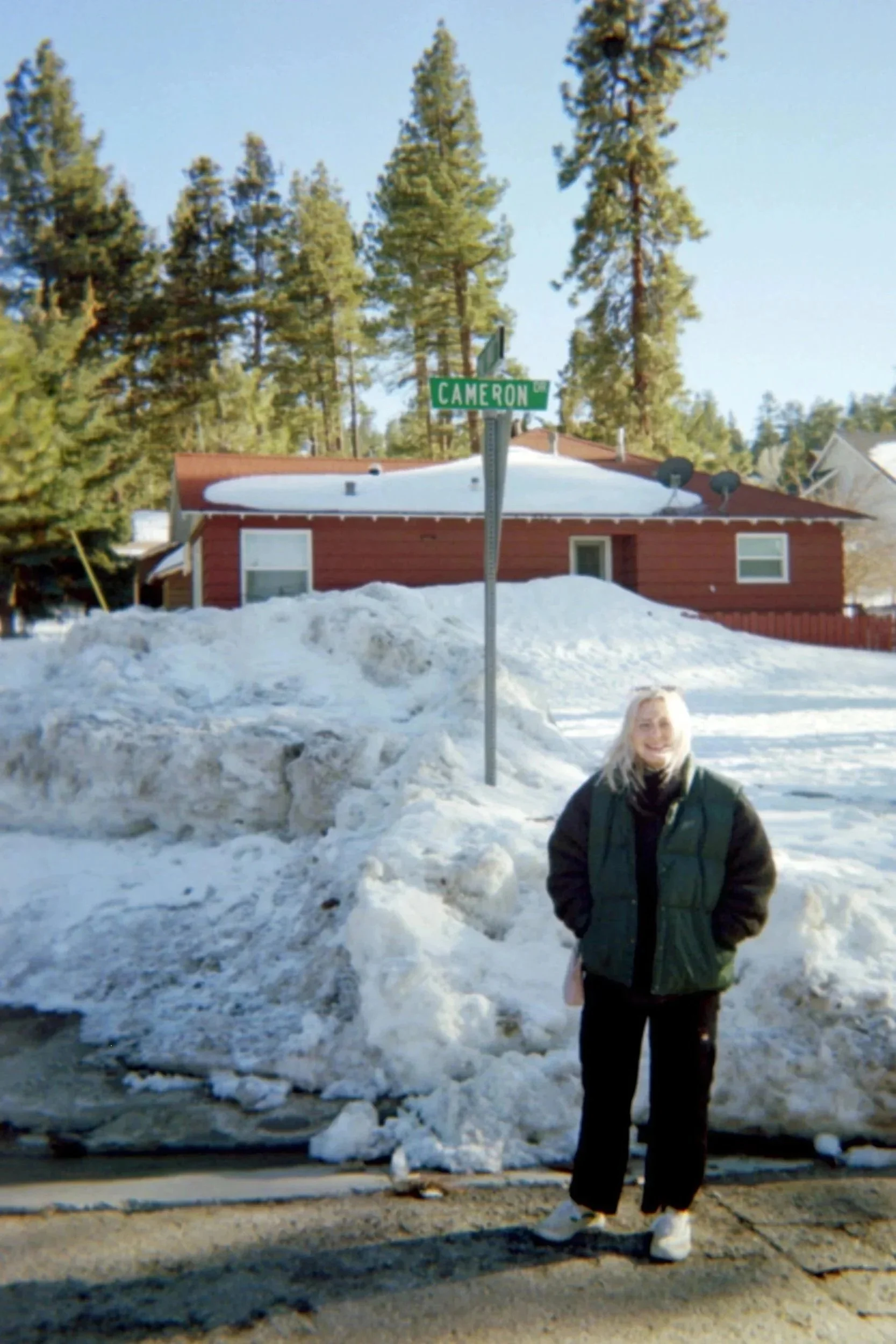 Woman standing on a snow-covered street corner at Cameron Street in a residential neighborhood with tall pine trees in the background, a red house, and a large snowbank.