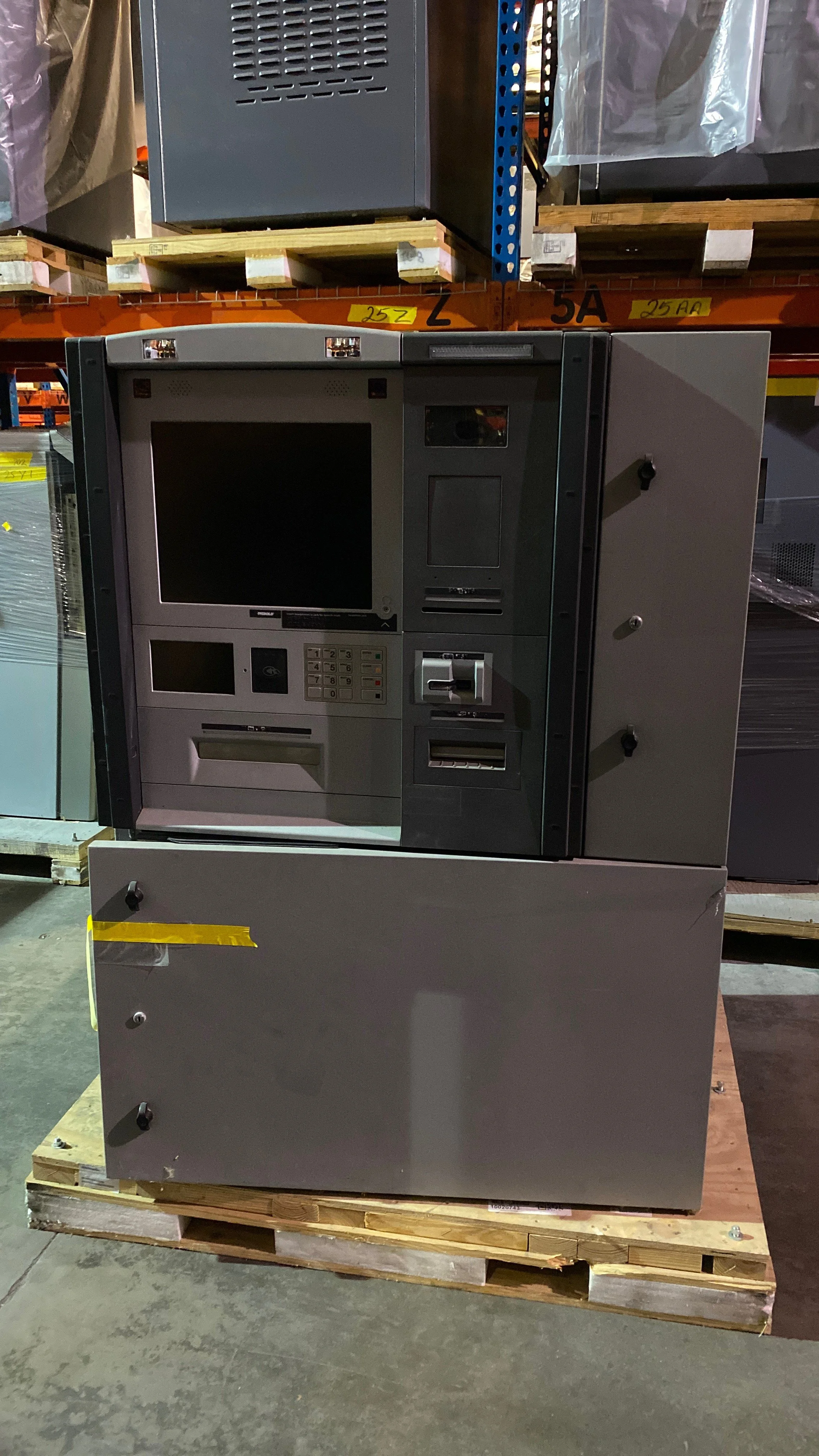 A grey ATM machine with various card slots, a keypad, and a screen, placed on a wooden pallet in a warehouse.