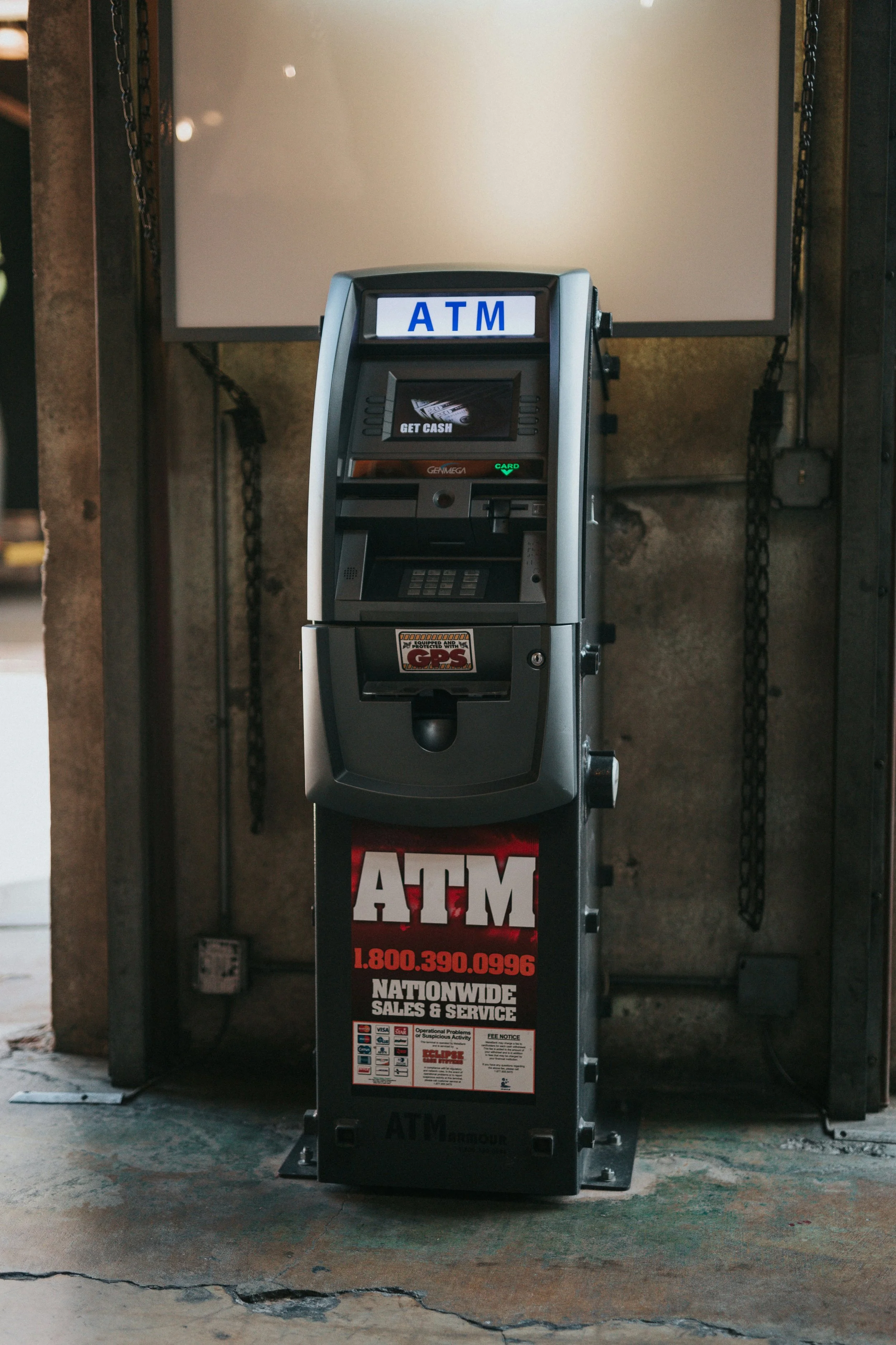 A black ATM machine standing on a concrete floor against a wall with chains. The ATM has a sign with blue lettering that reads "ATM" at the top and a sticker with red text advertising nationwide sales and service, along with a phone number.