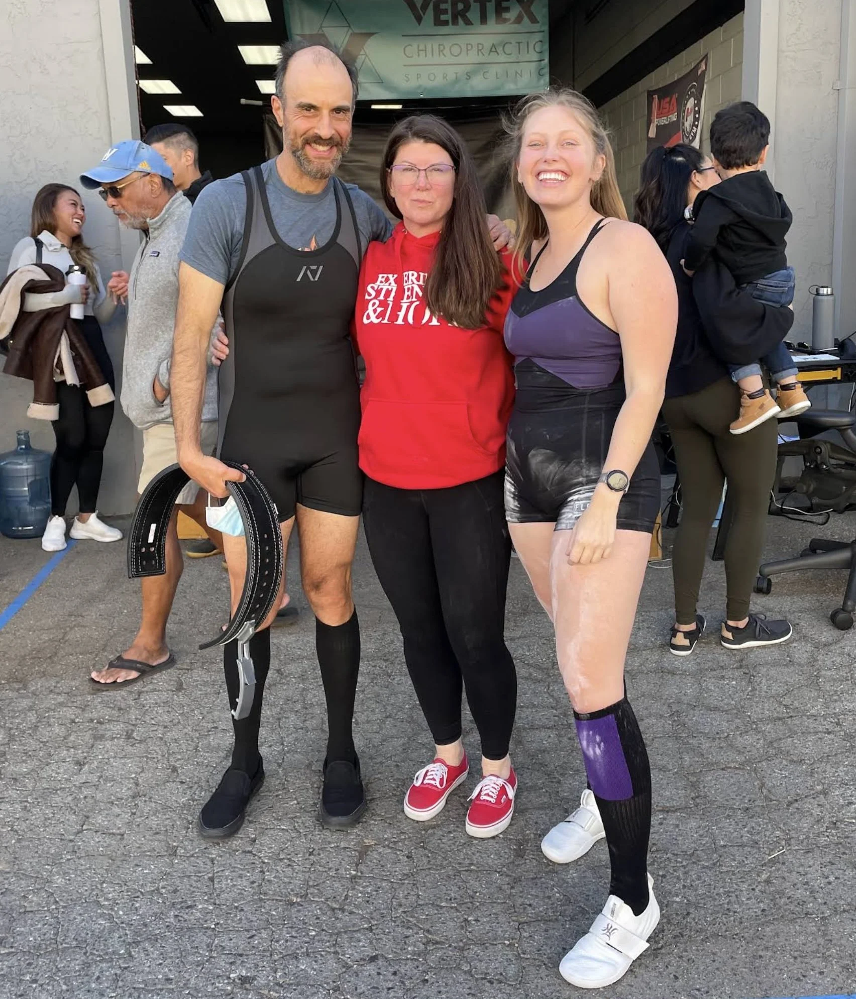 One man and two women standing outside a gym with their arms around each others' shoulders. Daune Turner is the woman in the middle and there are passers by in the background. The man to her right holds a back support belt. They are smiling.