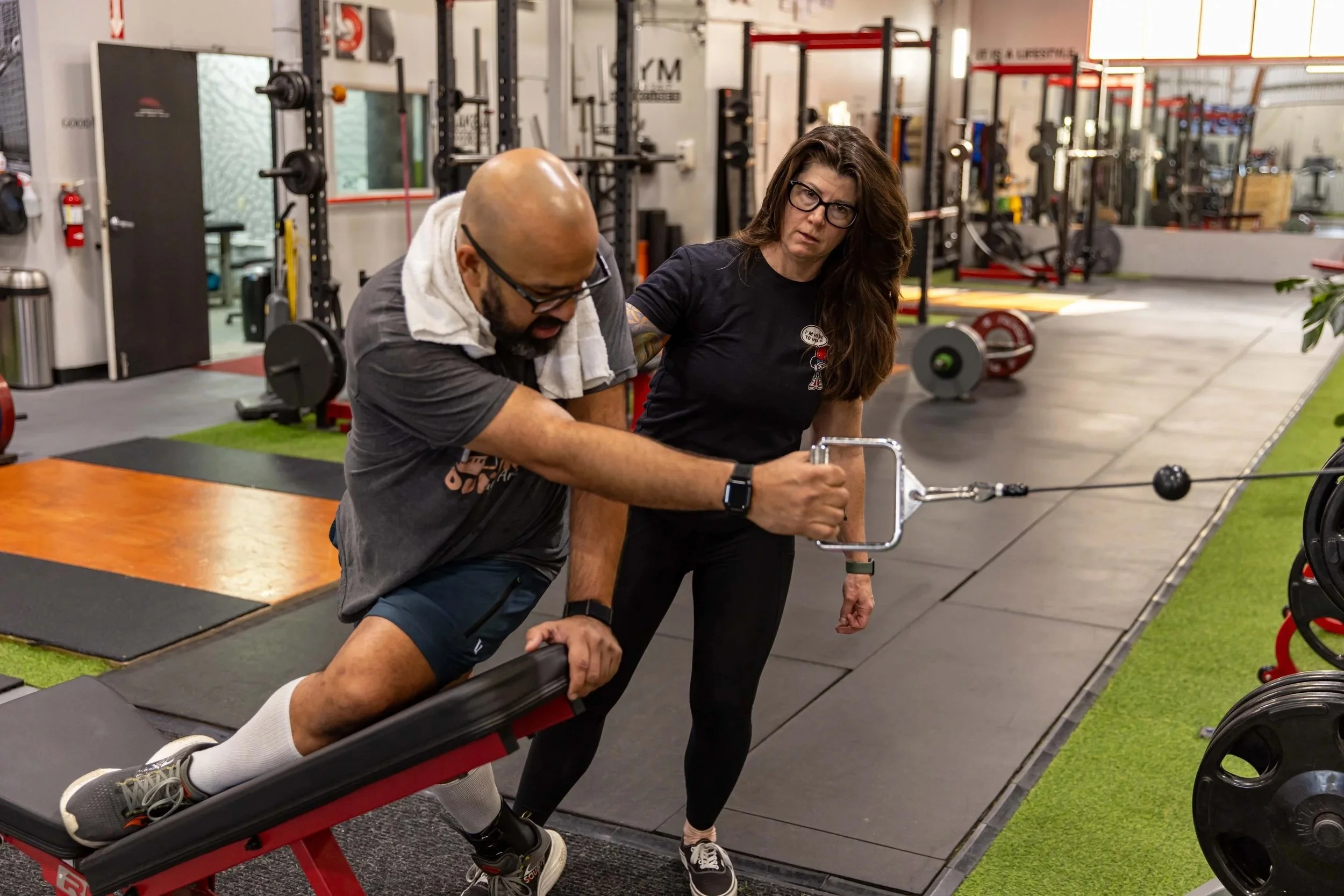 Vikas Chinnan completing a weighted spinal rotation exercise in a deep stretch to work on muscle rigidity. Daune Turner supervises in the background. They are in a gym with barbells and other weights in the background.