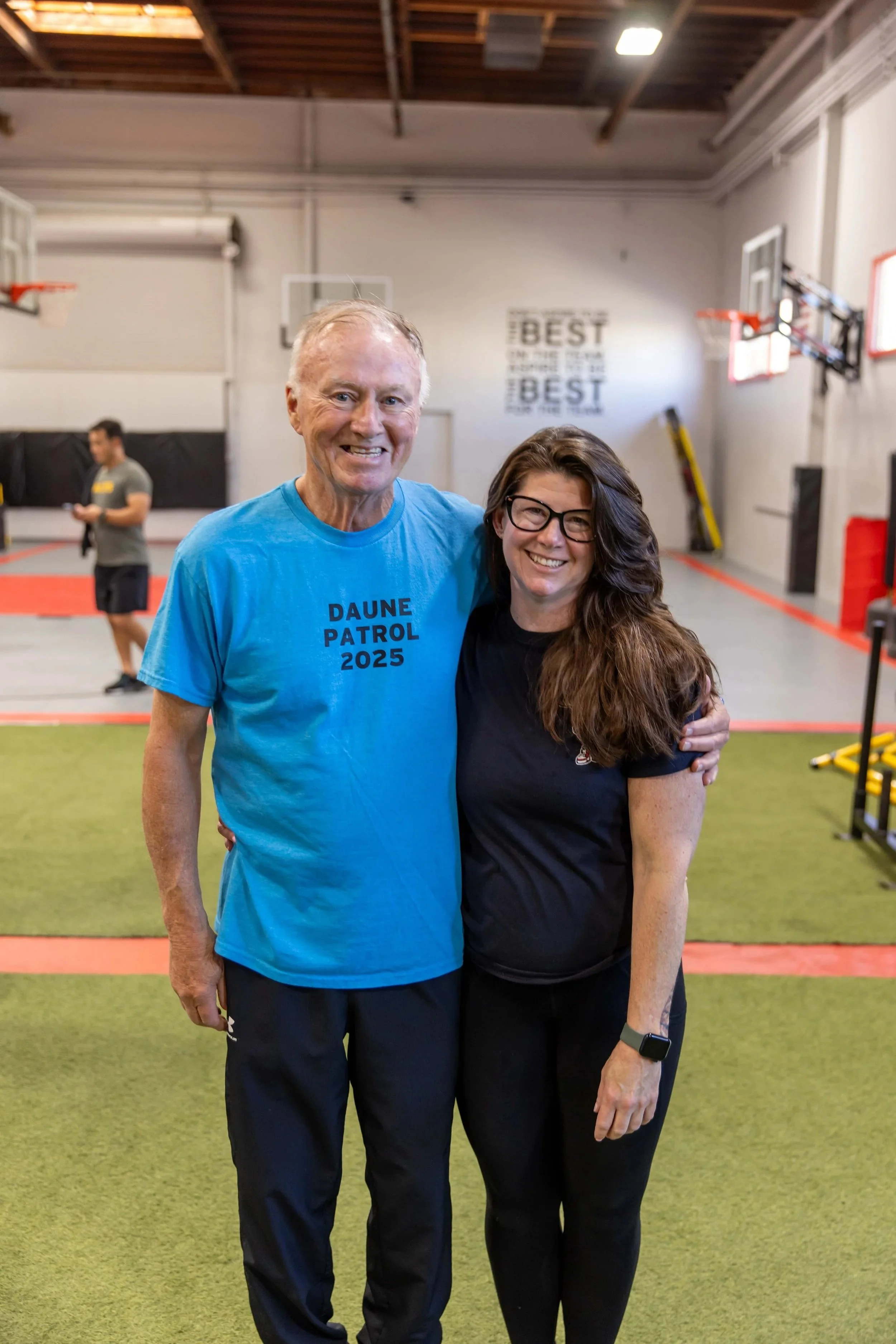 A cheerful older man with gray hair wearing a blue t-shirt that says “Daune Patrol 2025” next to Daune Turner, both smiling in a gymnasium. The background features a basketball hoop, safety equipment, and a motivational sign on the wall.
