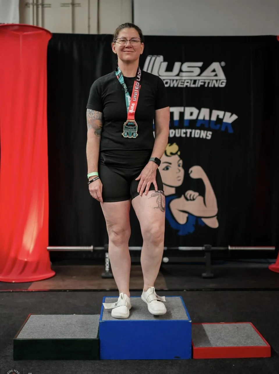 Daune Turner stands on a platform after achieving a California state squat record in the Ladies of Iron competition. She is wearing black exercise t-shirt and shorts and smiling.