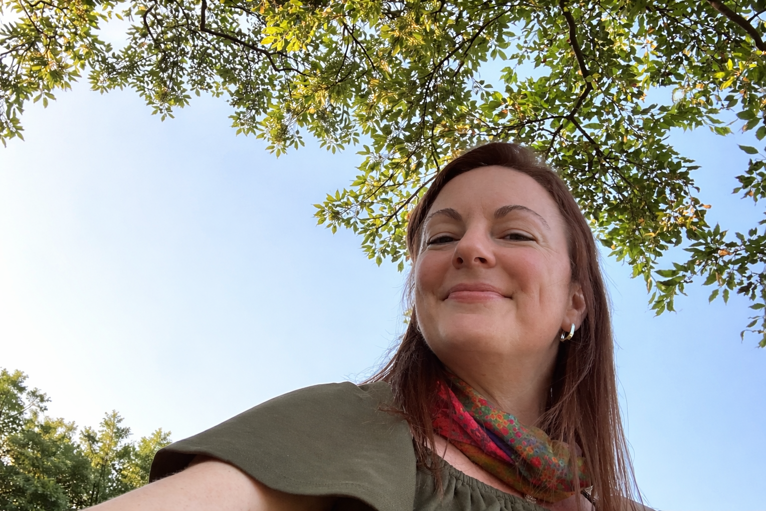 Smiling woman taking a selfie outdoors beneath leafy green trees, with sunlight filtering through the branches and a clear blue sky in the background.