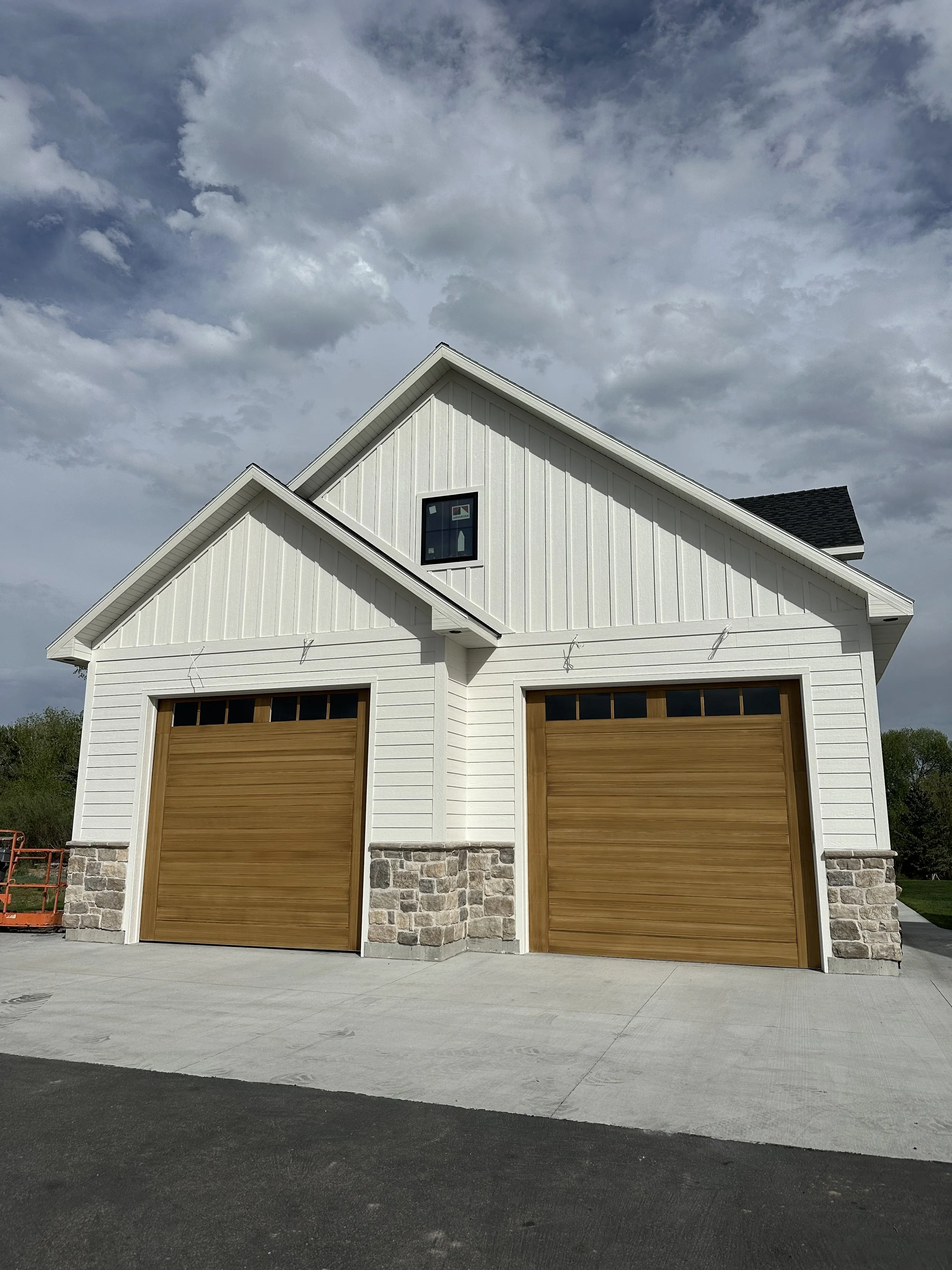 A modern white garage with two wooden doors, stone accents at the bottom, and a small window in the upper part, set against a cloudy sky.